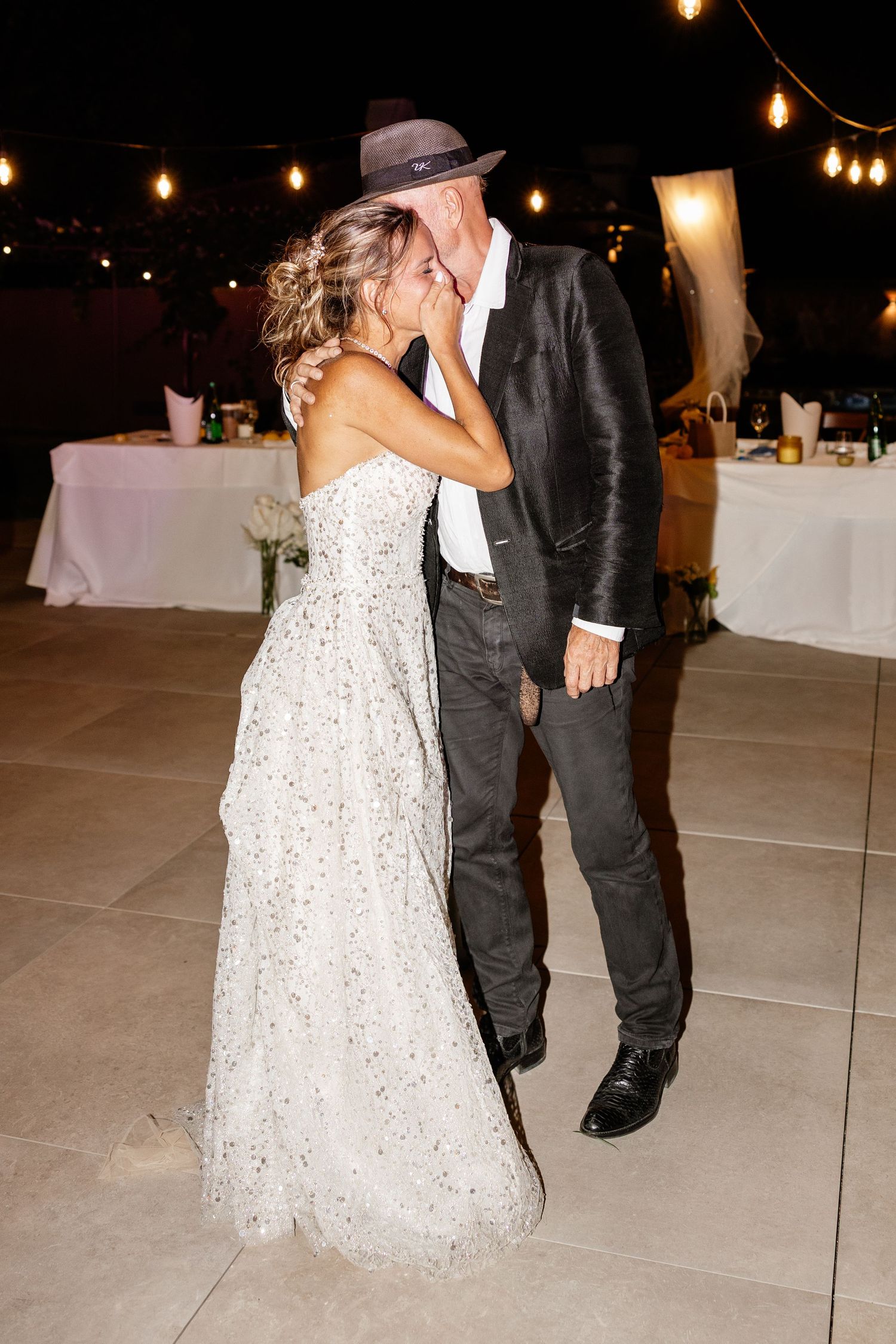Bride and groom sharing a kiss during evening outdoor wedding reception dance under string lights.