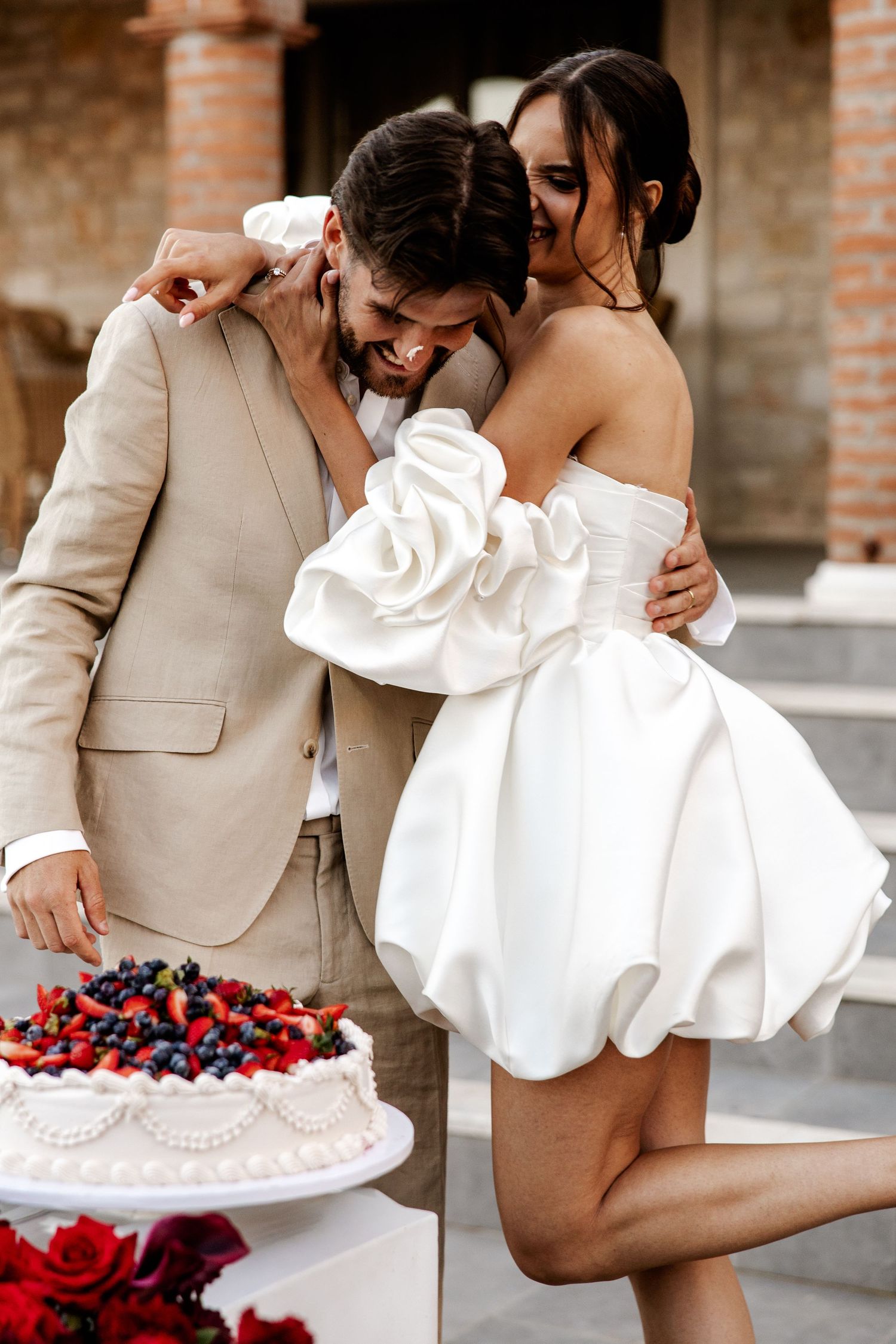 Bride in white mini dress embraces groom in beige suit beside decorated wedding cake with fresh berries.
