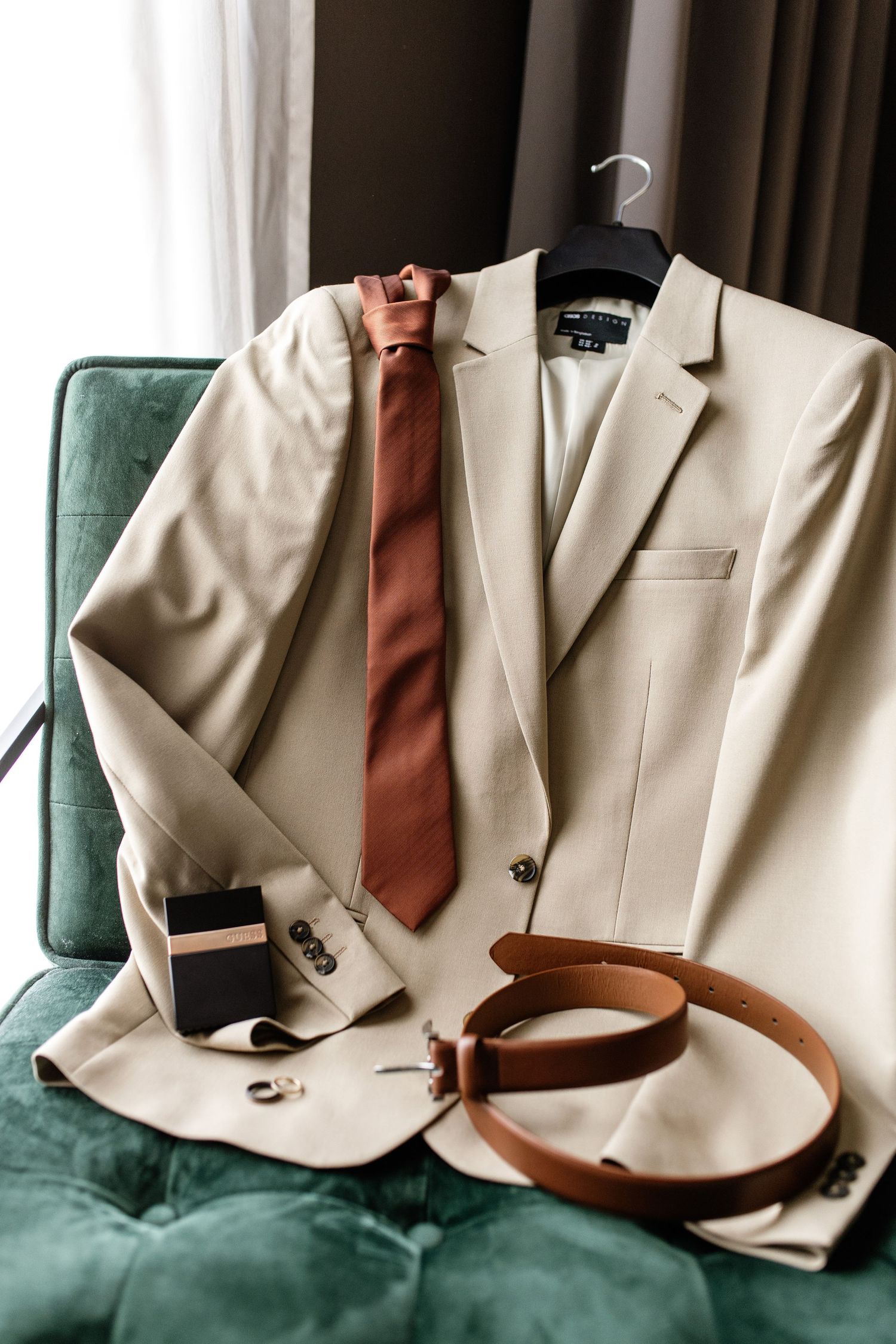 Elegant groom's wedding accessories including beige suit, brown tie, leather belt, cufflinks, and cologne on green velvet chair.