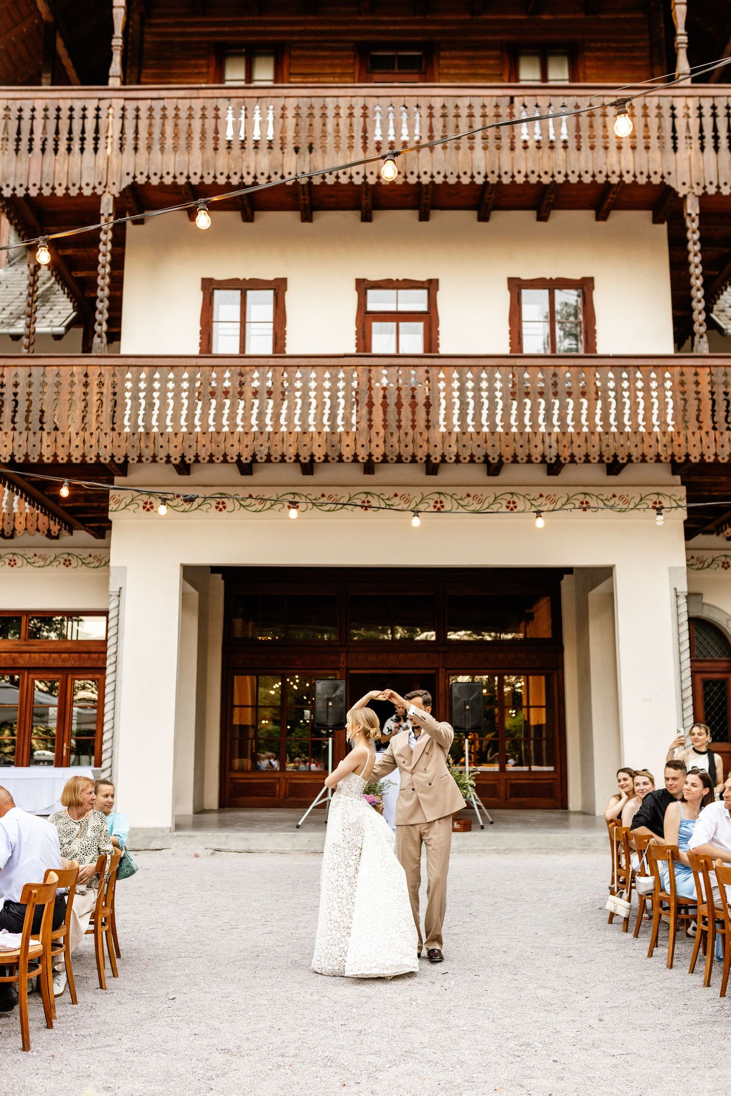 Bride and groom share first dance outside elegant venue with wooden balcony and string lights at outdoor wedding.