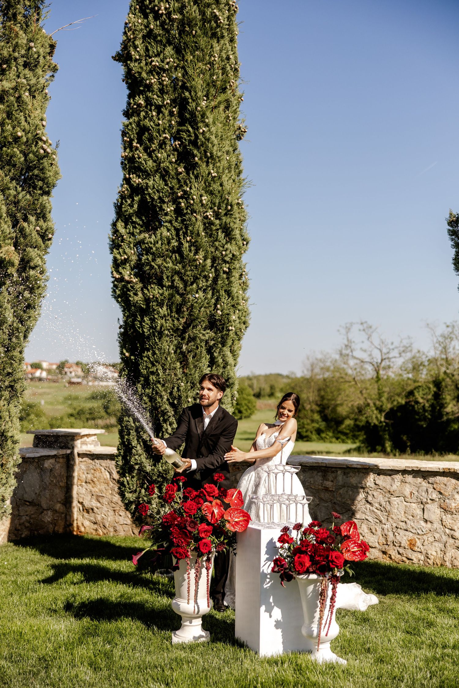 Bride and groom celebrating with champagne outdoors, surrounded by red roses and tall cypress trees on a sunny wedding day.