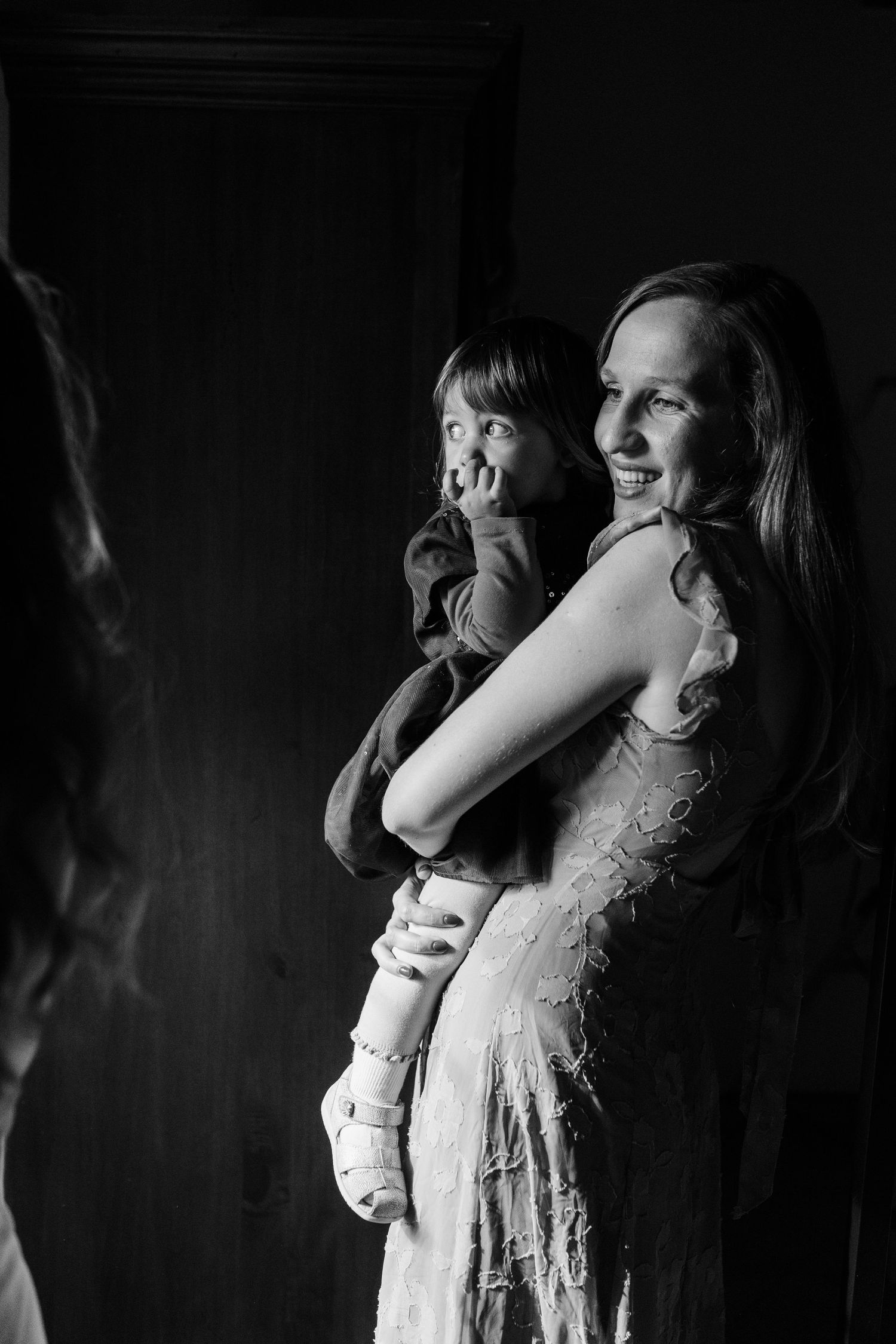 Smiling woman in white dress holds young child at wedding celebration in black and white.