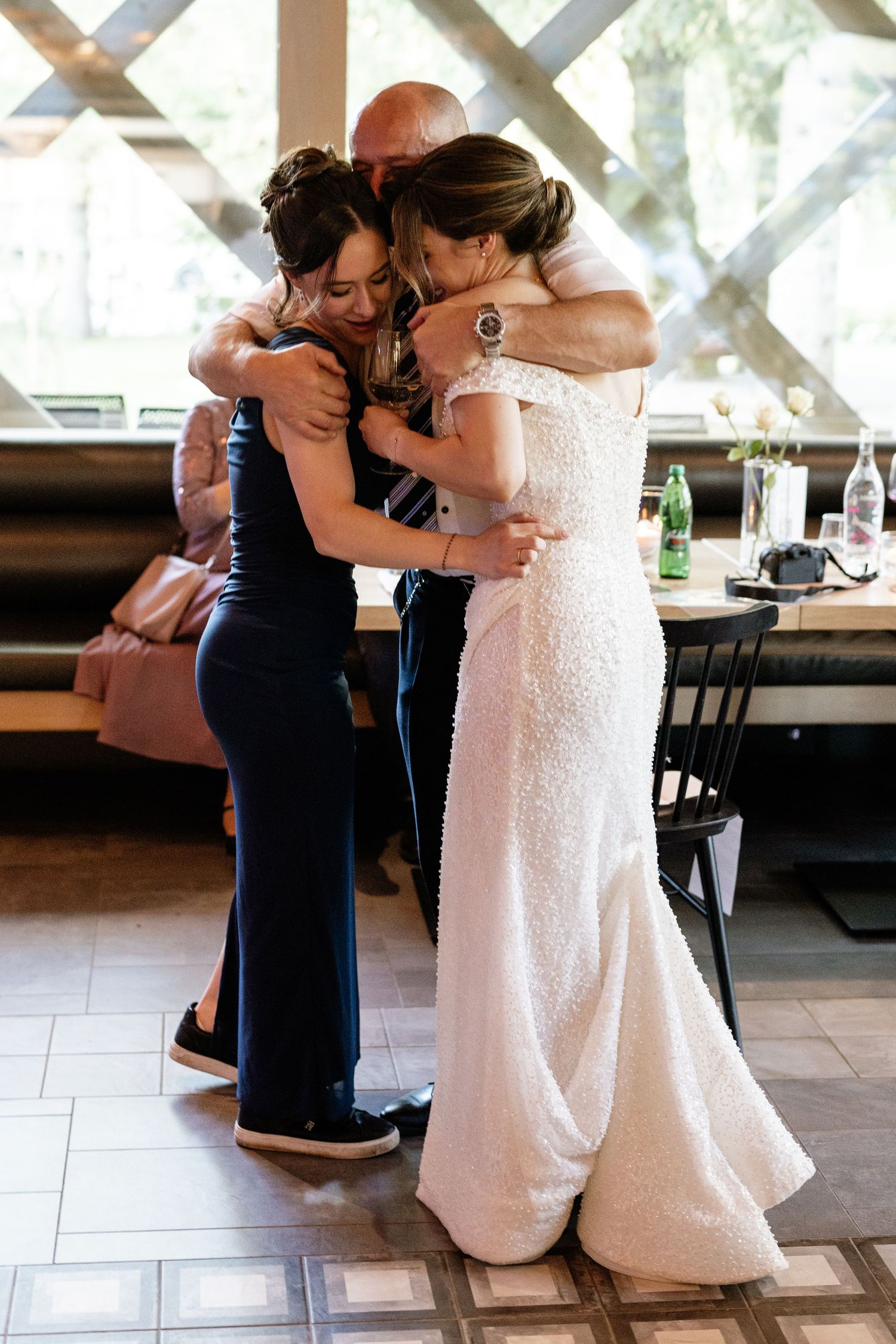 Bride in white lace gown shares emotional group hug with two guests at wedding reception indoors.
