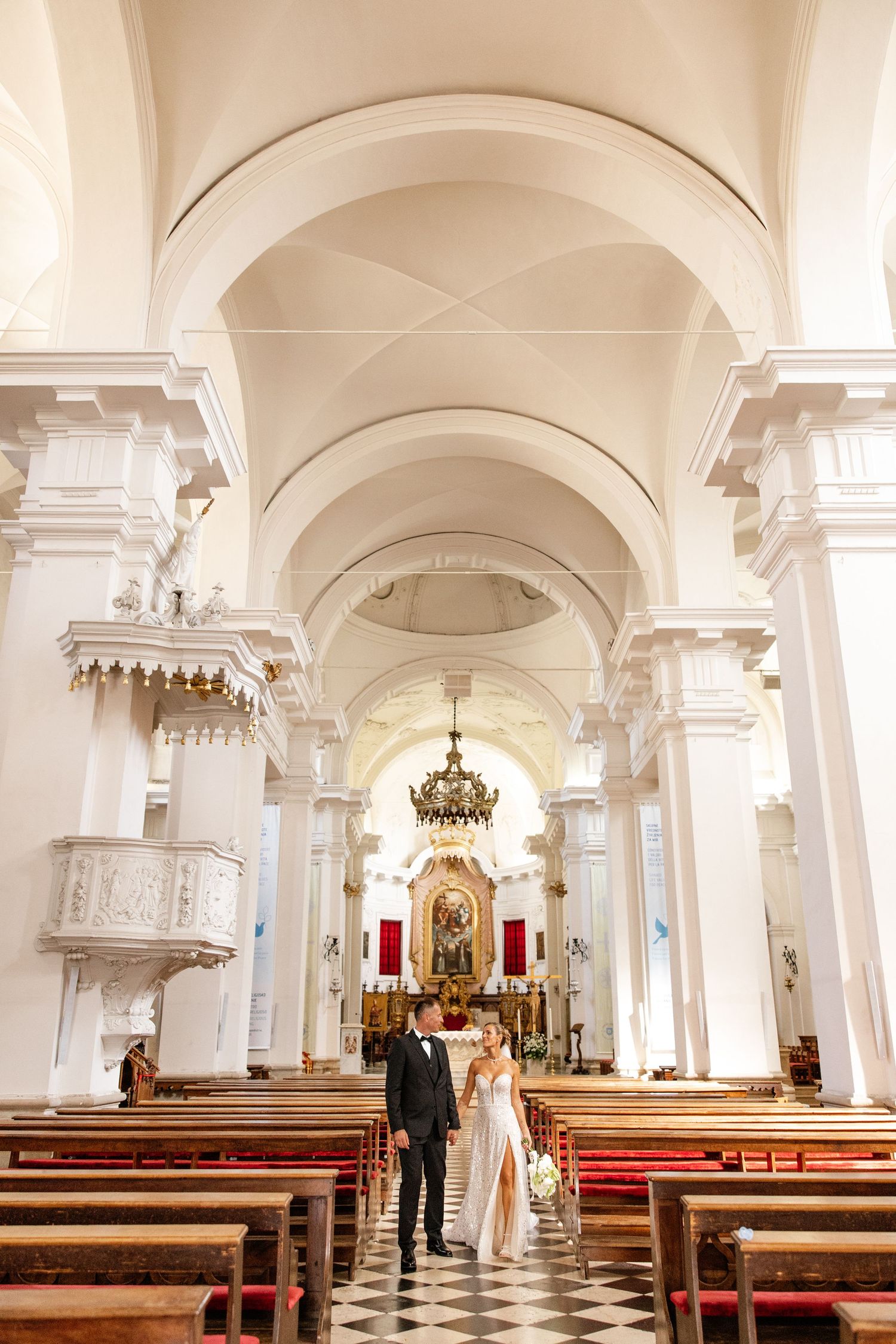 Bride and groom walking down the aisle of an elegant white baroque church with arched ceilings and wooden pews.