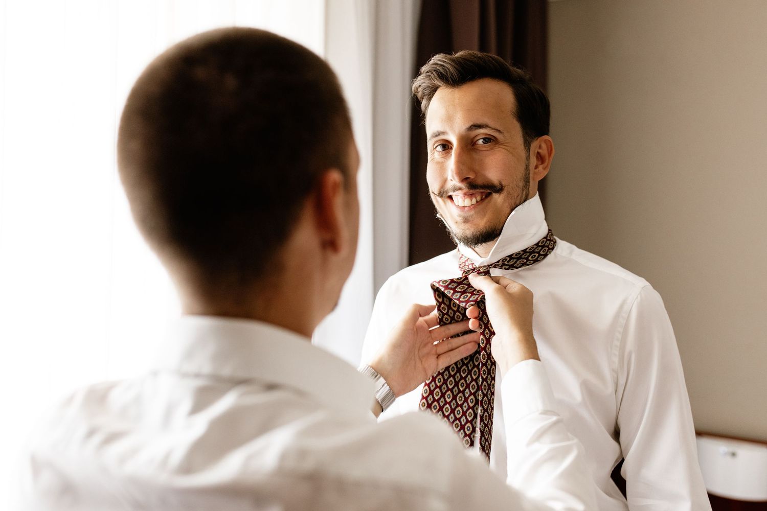 A smiling groom adjusts his patterned tie with help from a groomsman in a white shirt before the wedding.