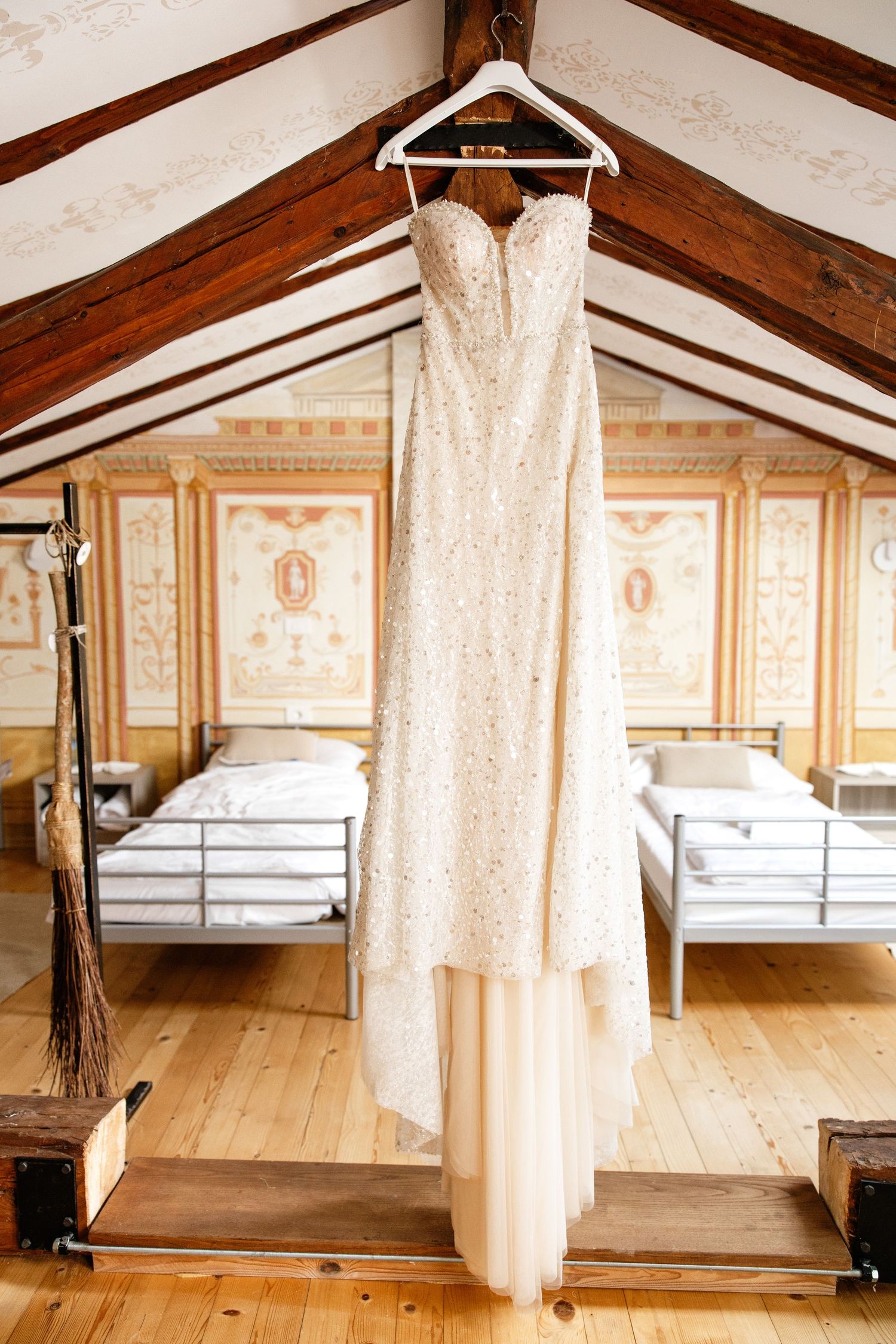 Elegant beaded strapless wedding gown hanging on white hanger in rustic attic room with ornate wall murals.