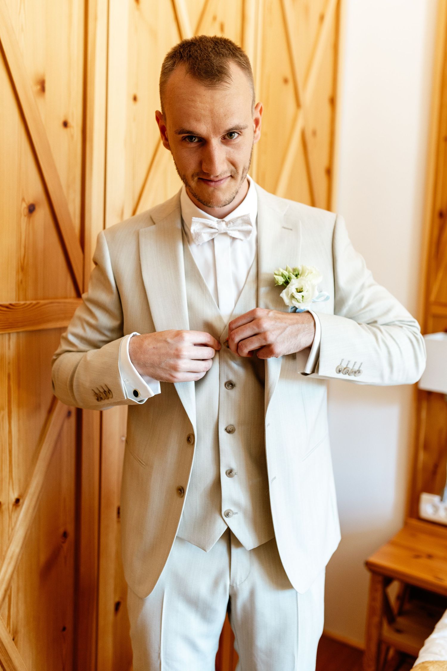 Groom in beige suit with bow tie and boutonniere buttoning jacket in wooden interior before wedding.