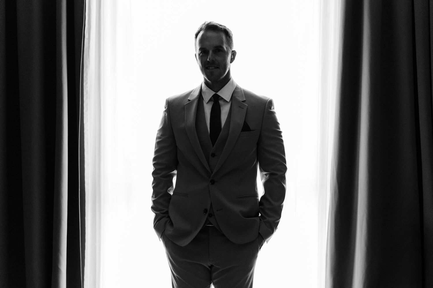 Black and white silhouette of a man in a tailored suit standing between curtains with hands in pockets.