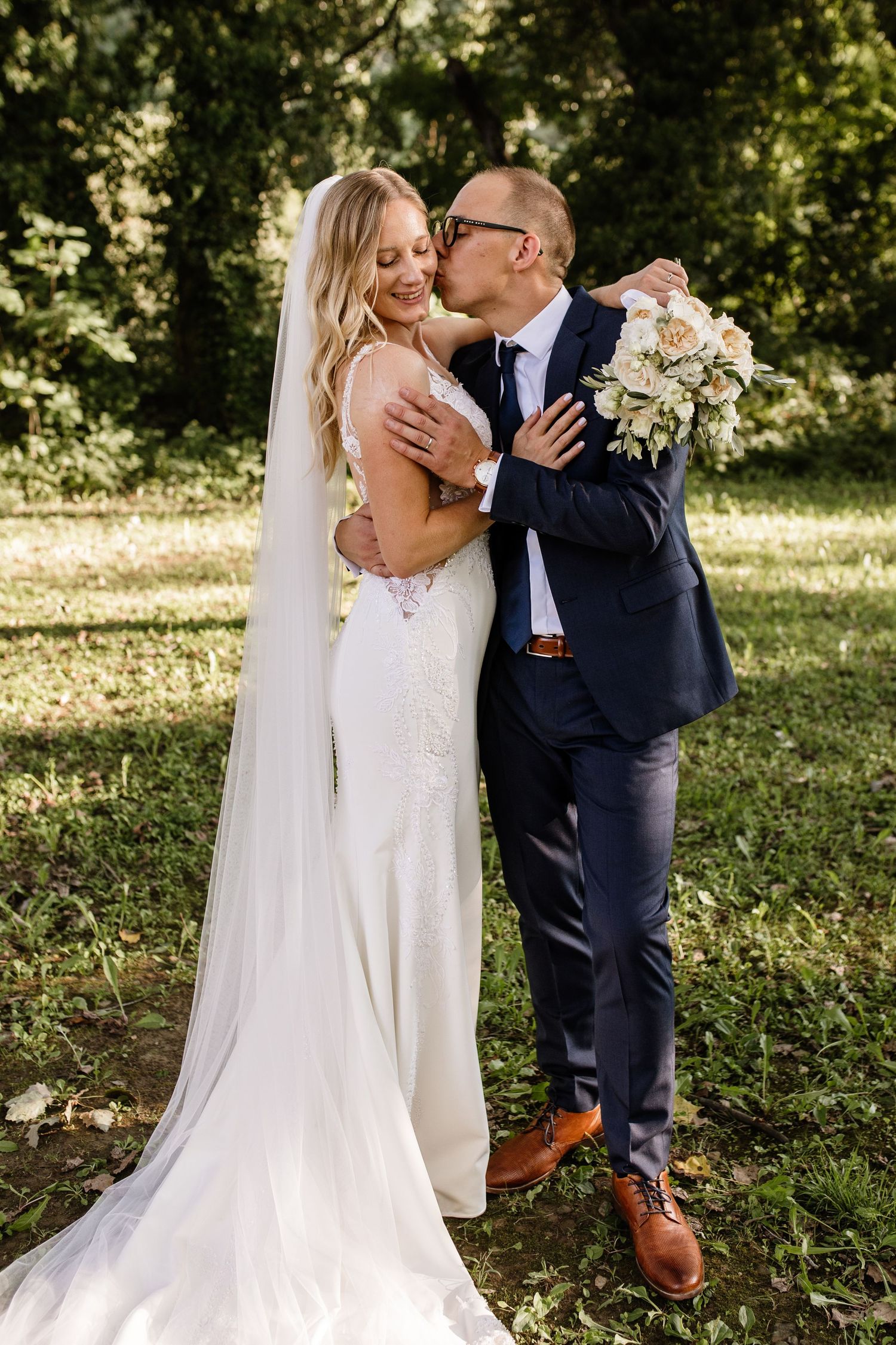 Groom in navy suit kisses bride in white gown and veil while holding bouquet outdoors in lush green garden.