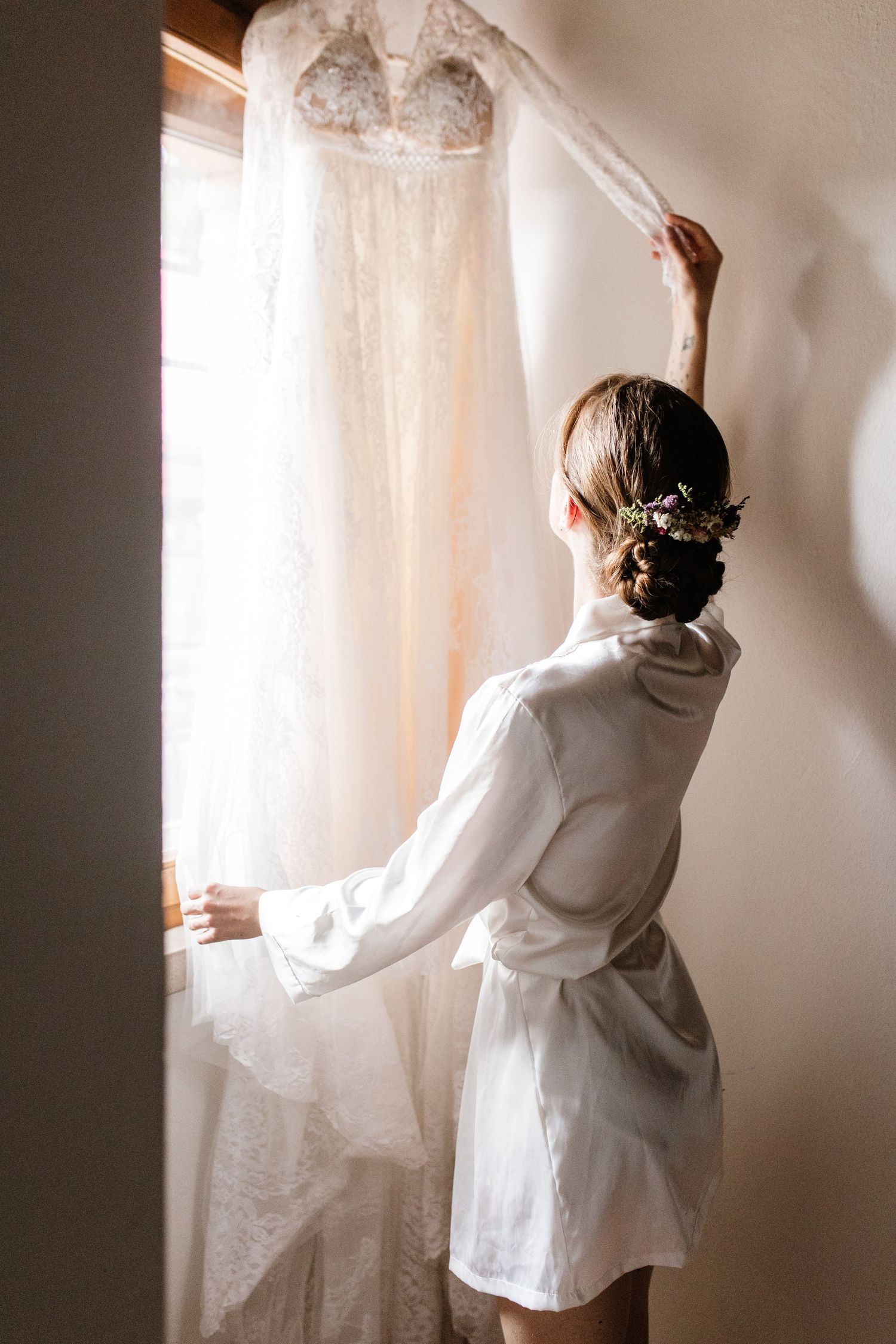 Bride in white robe reaches for lace wedding dress hanging by sunlit window.