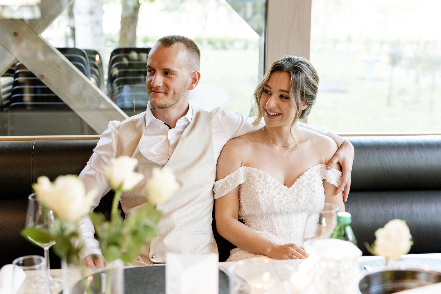 Happy couple in wedding attire seated together at a reception table with white roses in the foreground.