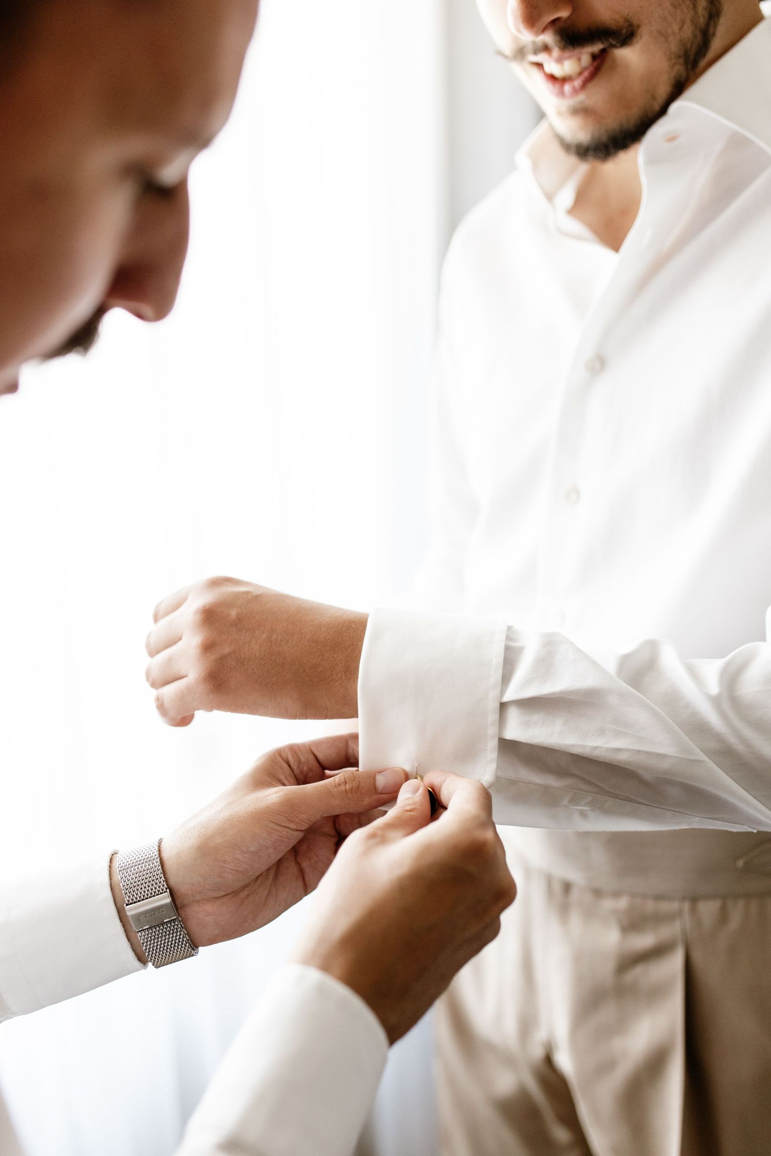 A man helping another man fasten his cufflinks on a white dress shirt before a formal event.