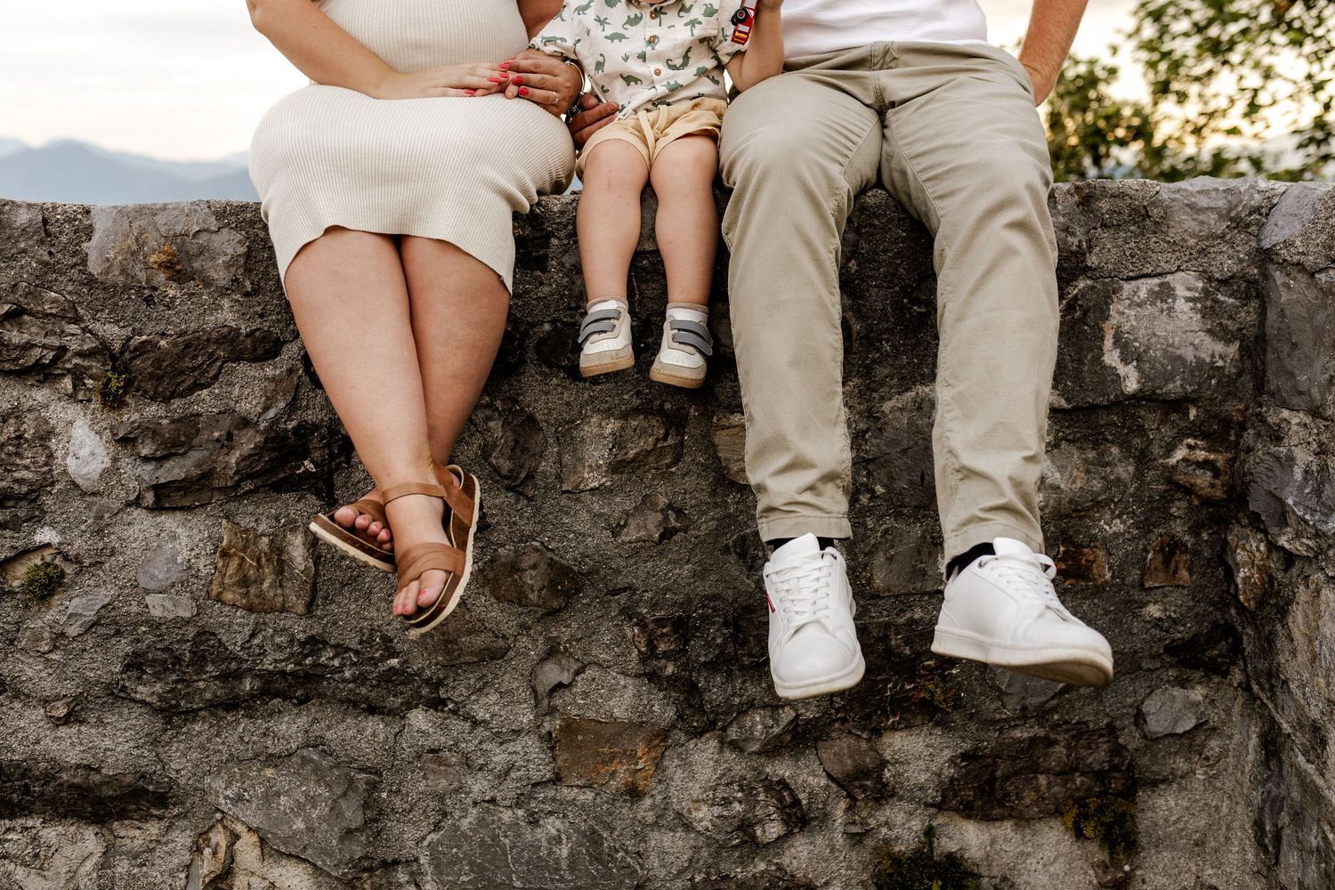 Family sitting on a stone wall, legs dangling, wearing casual shoes, with a child between two adults outdoors.