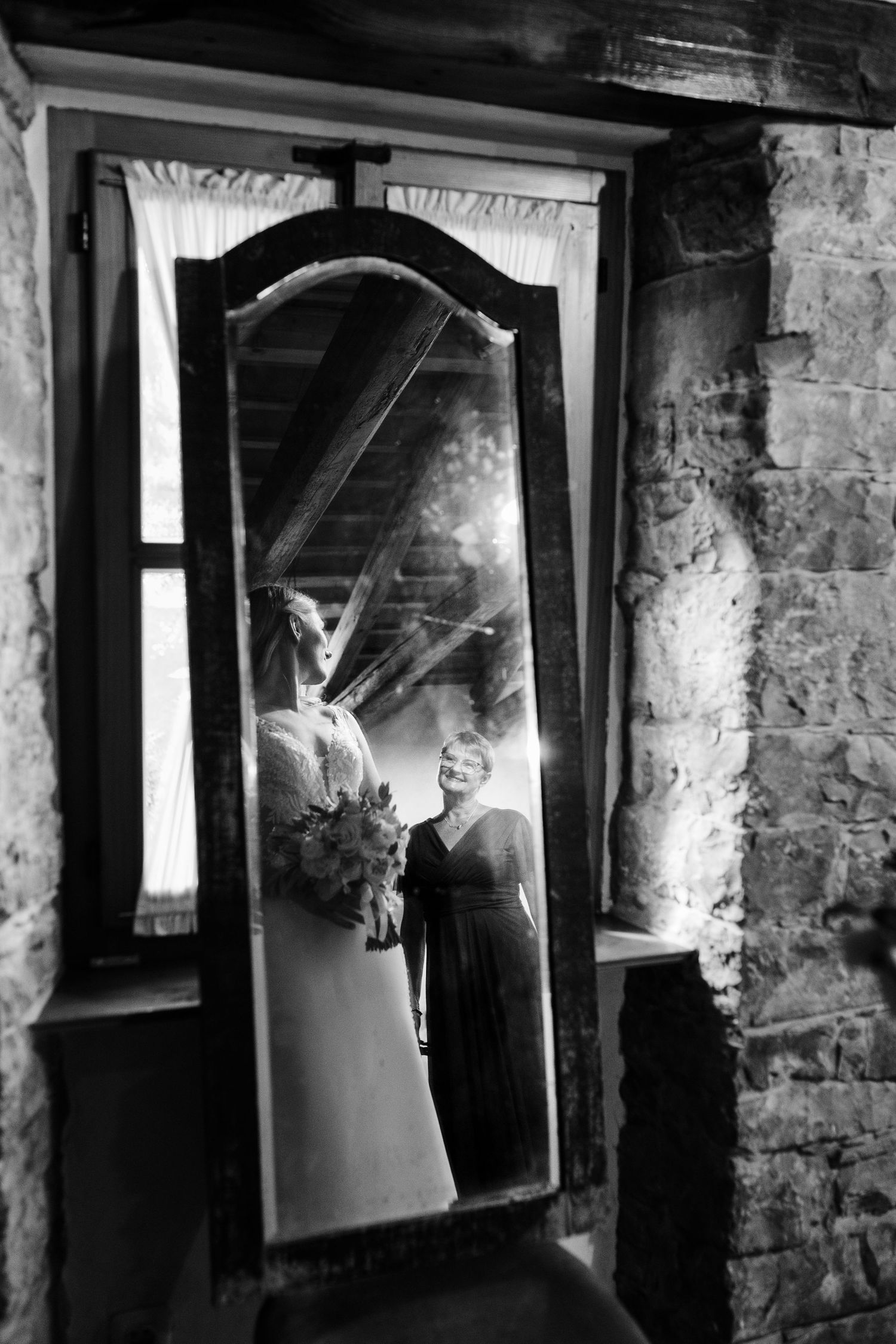 Black and white photo of a smiling bridesmaid holding a bouquet reflected in a vintage floor mirror in a rustic stone room.