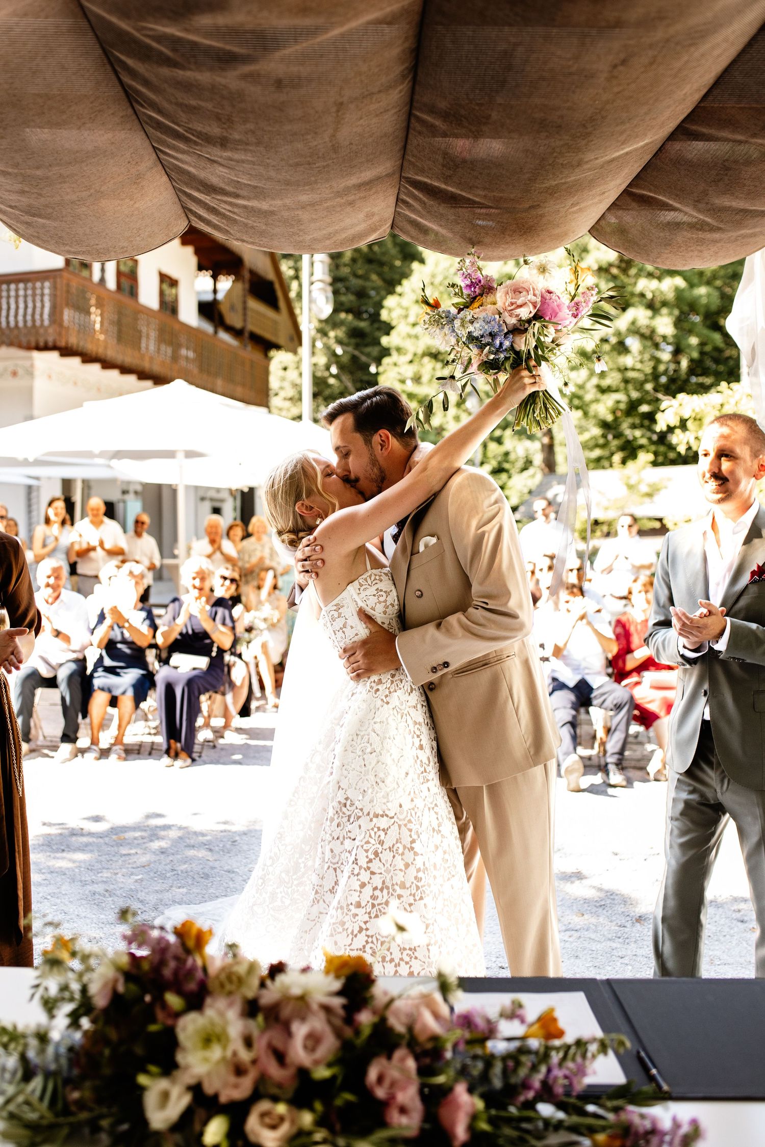 Bride and groom share first kiss at outdoor wedding ceremony, bride holding colorful bouquet aloft.