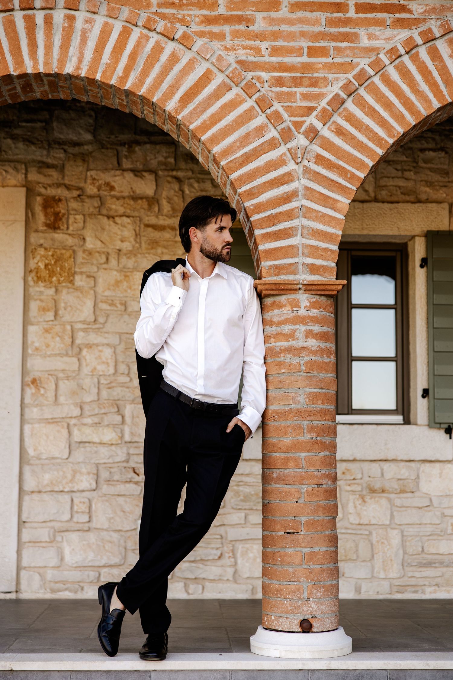 Stylish groom in white shirt and black trousers leaning against a brick column under a rustic stone archway.