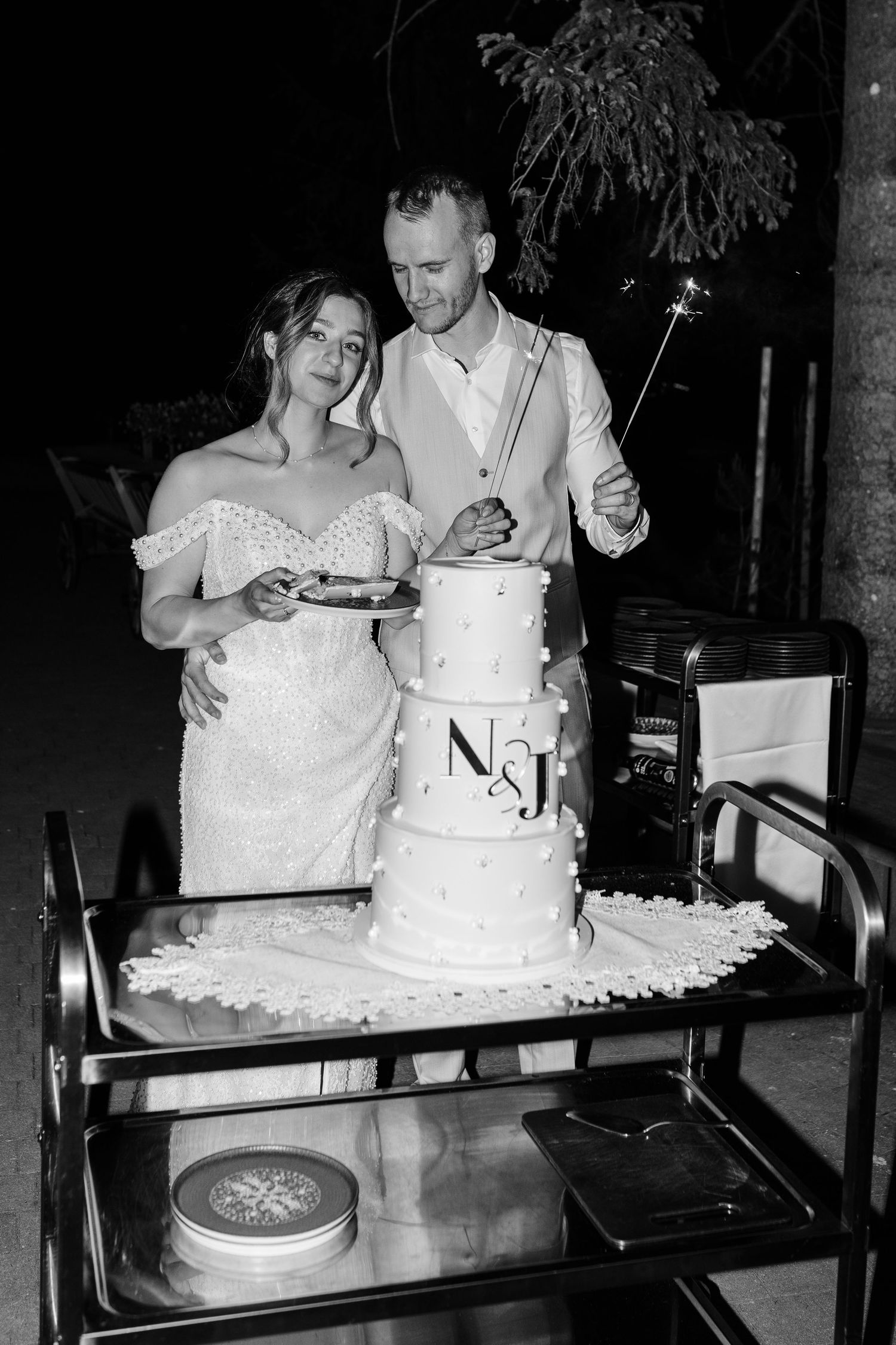 Bride and groom cutting their three-tier wedding cake monogrammed with N&J at an outdoor evening reception.