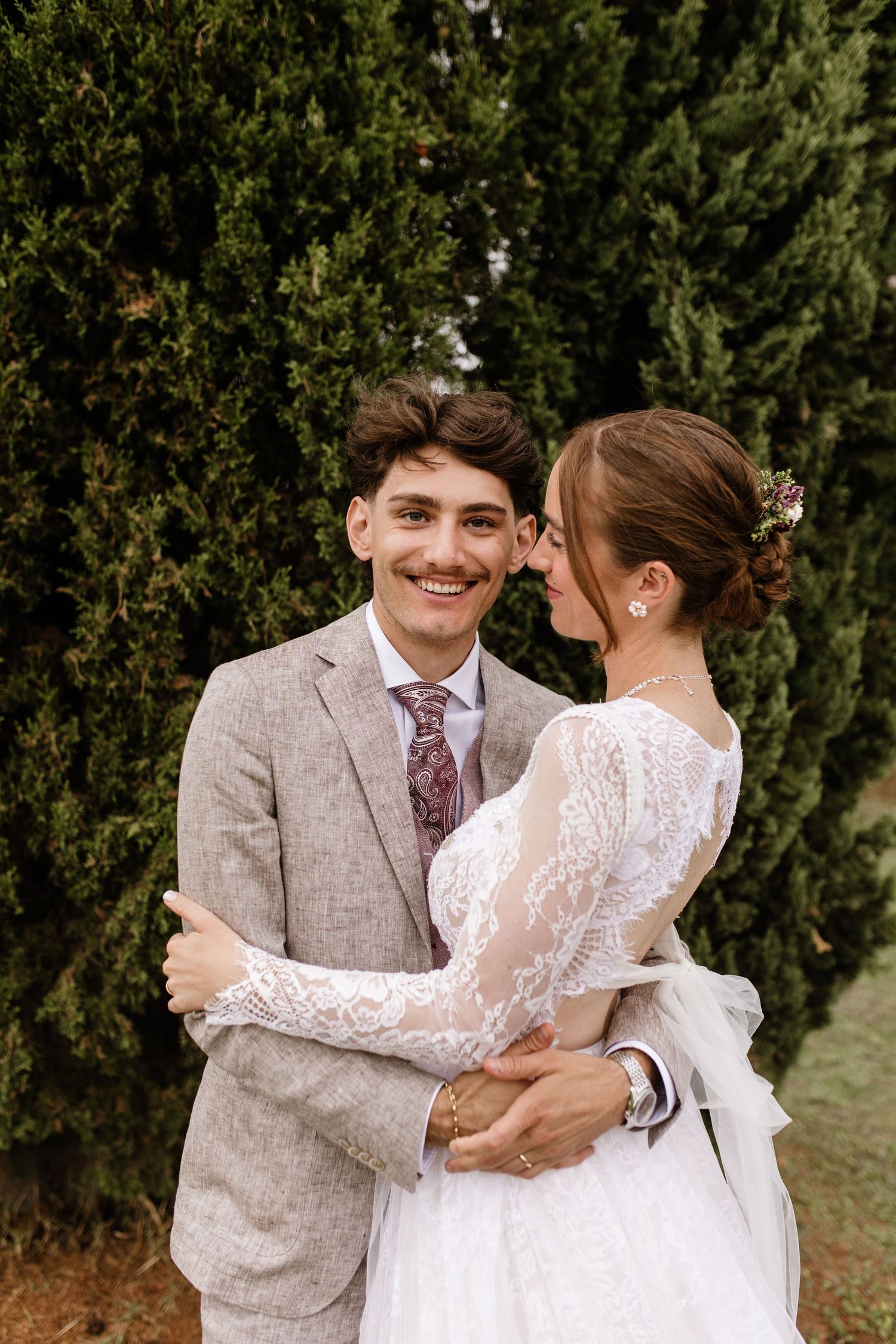 Smiling groom in gray suit embraces bride in lace dress before tall cypress trees.