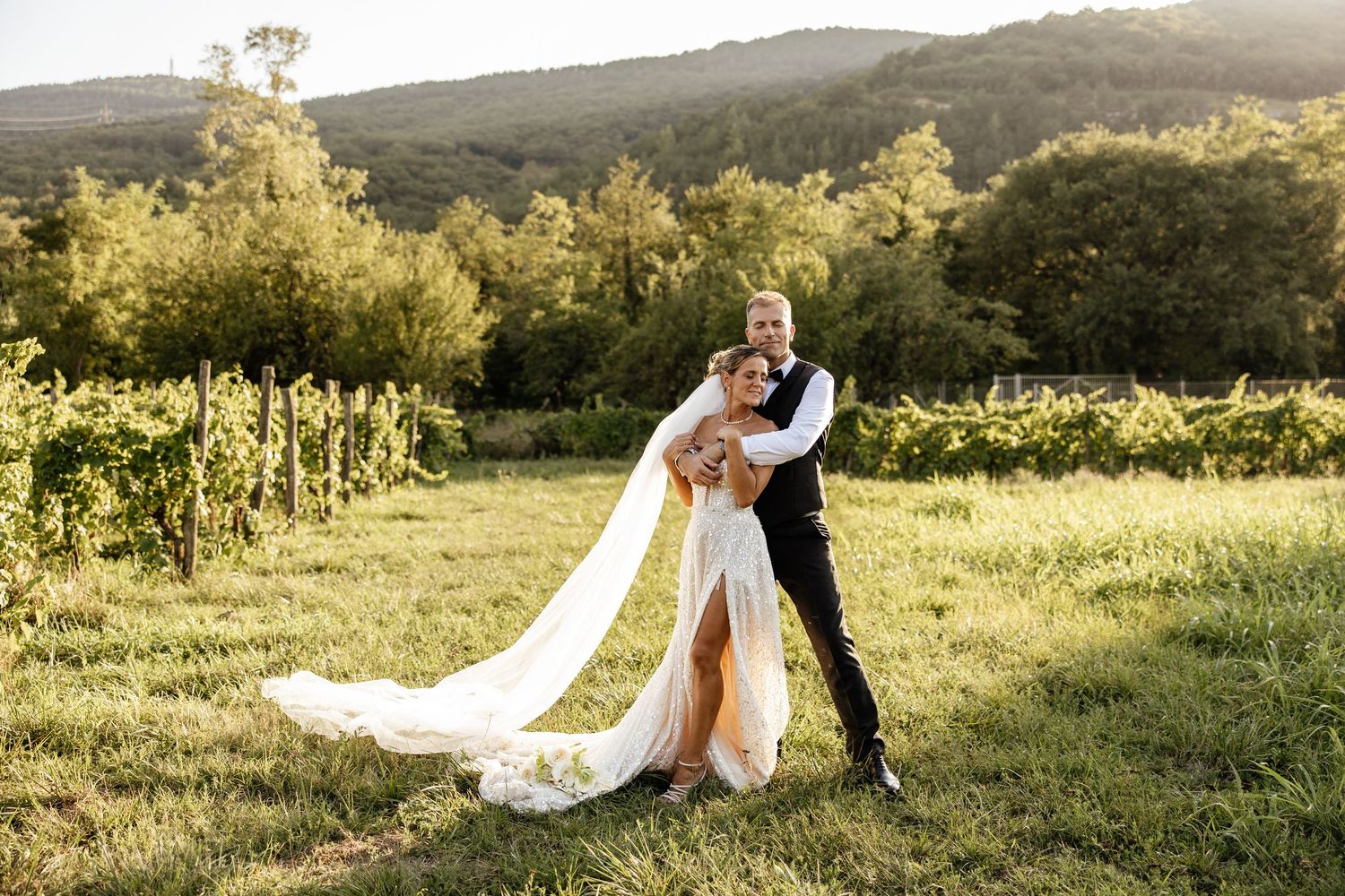 A couple embracing in a sunlit vineyard, bride in a flowing white gown with long veil, groom in black suit.