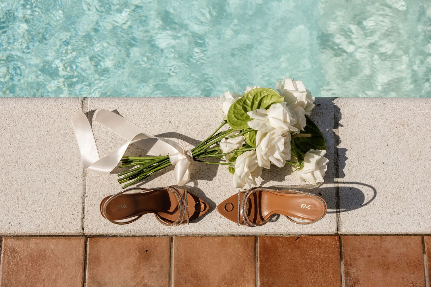 Brown strappy heels and white rose bouquet with ribbon on pool edge in sunlight.