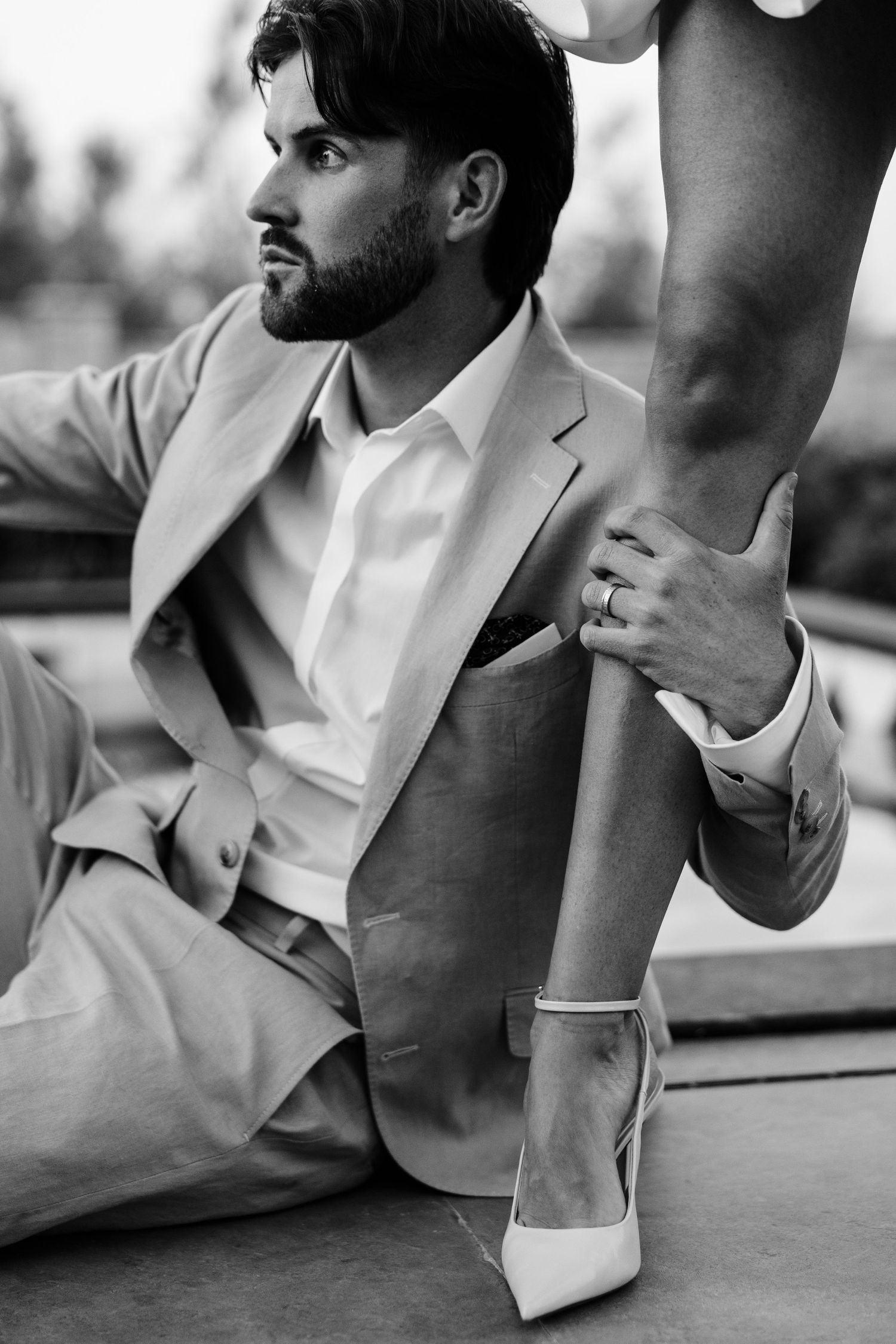 Black and white photo of a man in a suit sitting while holding a womans leg wearing white heels.