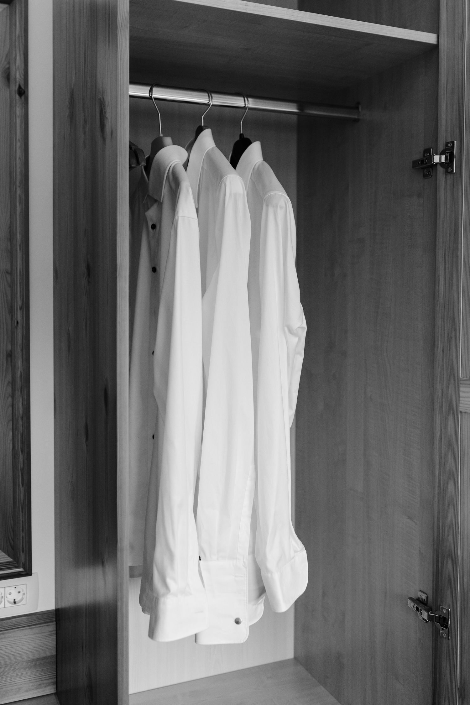 Three white dress shirts hanging neatly on hangers inside a wooden wardrobe closet, black and white photo.