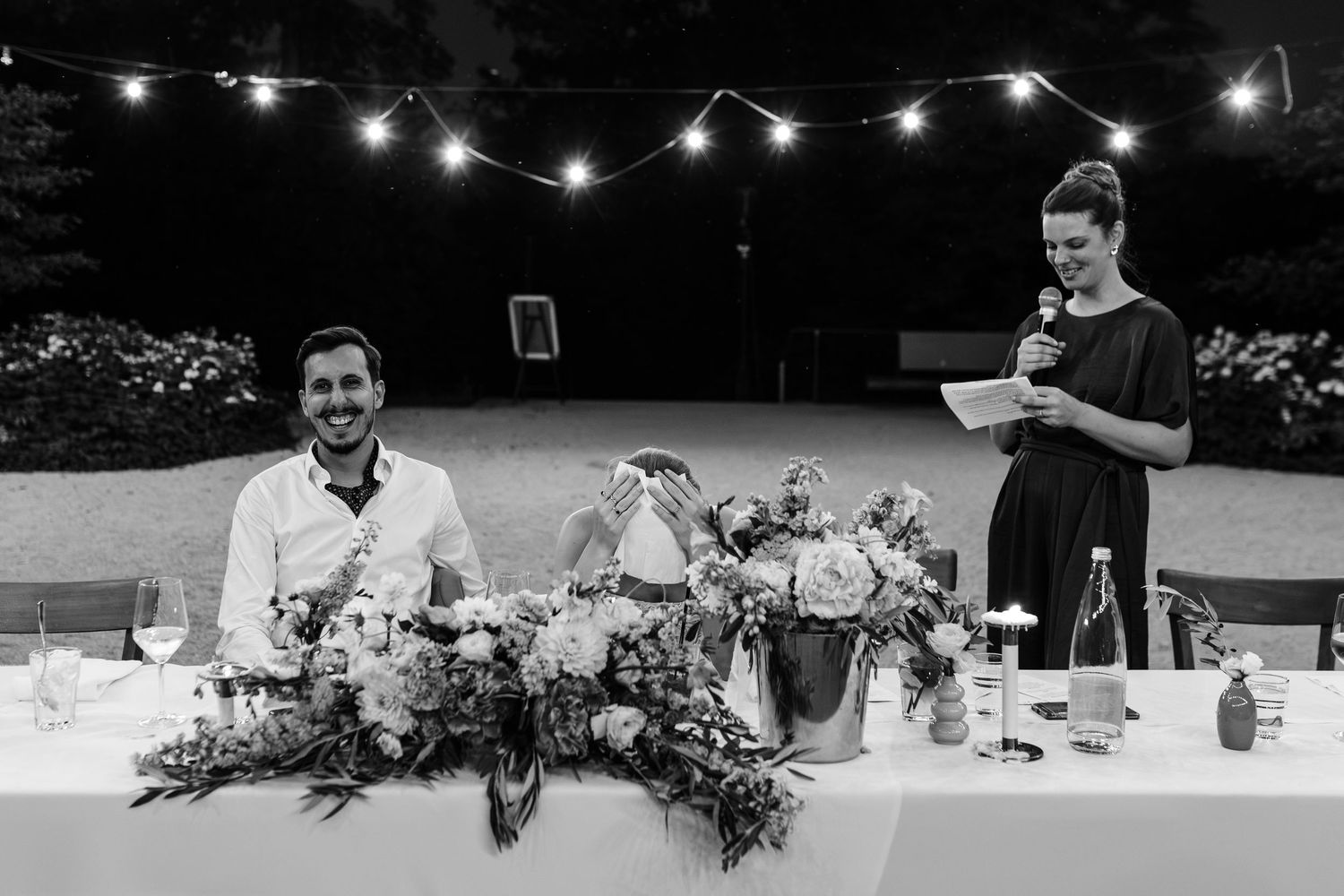 Black and white wedding reception photo with couple at floral table, woman giving speech under string lights outdoors.