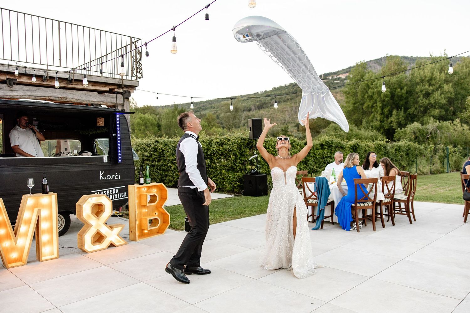 Bride tosses her veil as couple celebrates outdoors at night wedding reception with lit marquee letters.