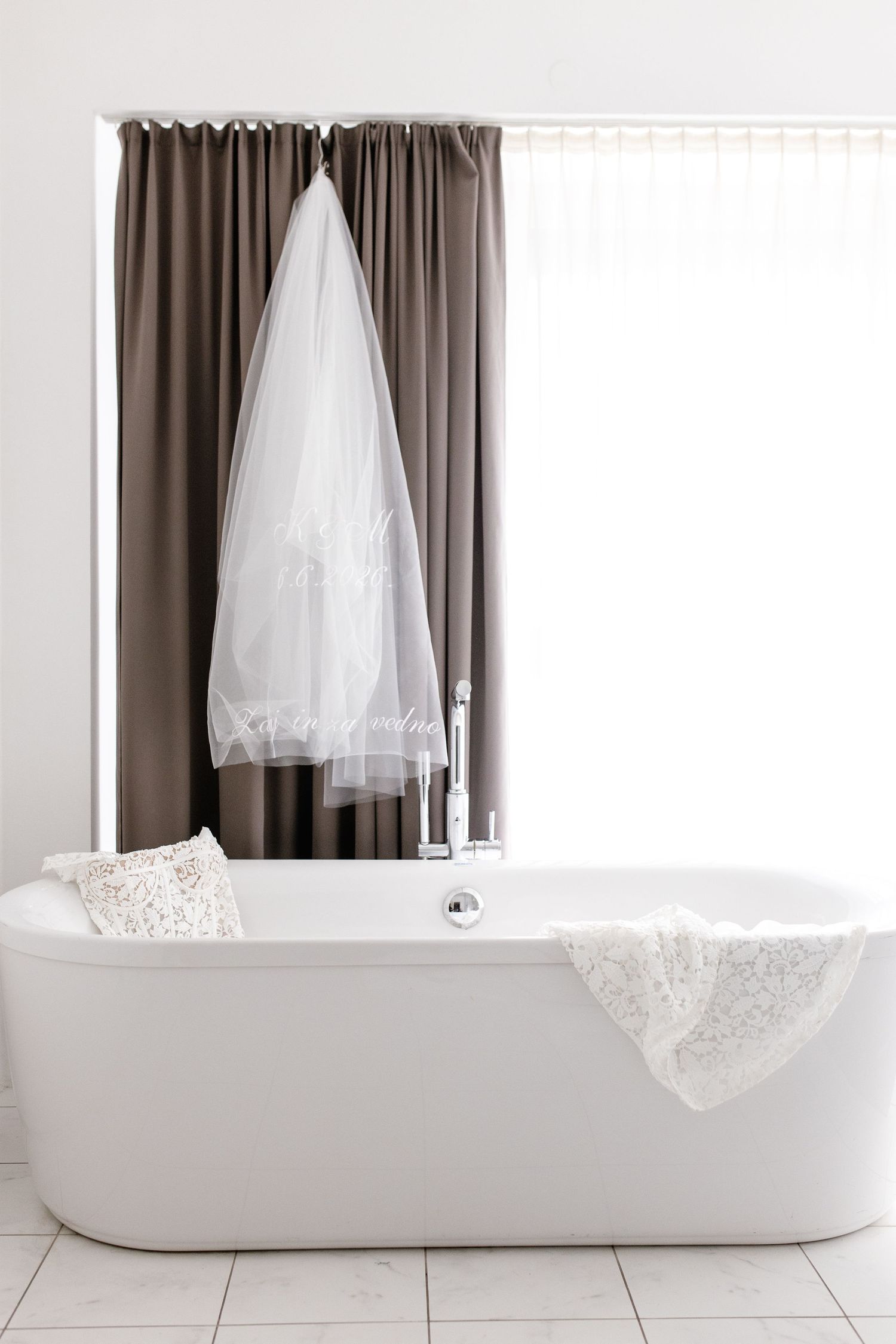 Elegant white freestanding bathtub with wedding veil hanging on brown curtains in a bright modern bathroom.