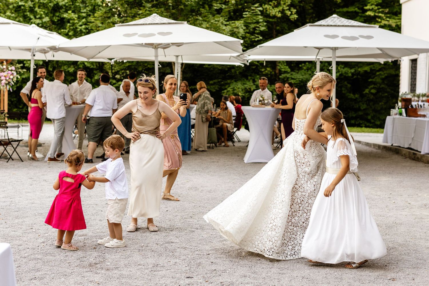 A bride in a white gown dances with children and guests at an outdoor wedding reception under white umbrellas.