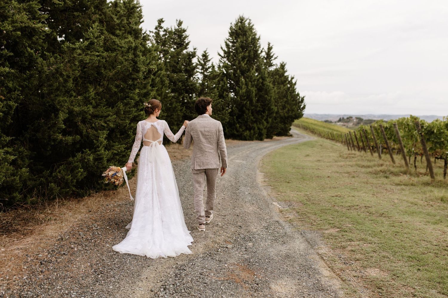 Bride and groom walking hand in hand down a gravel path lined with trees at a vineyard wedding.