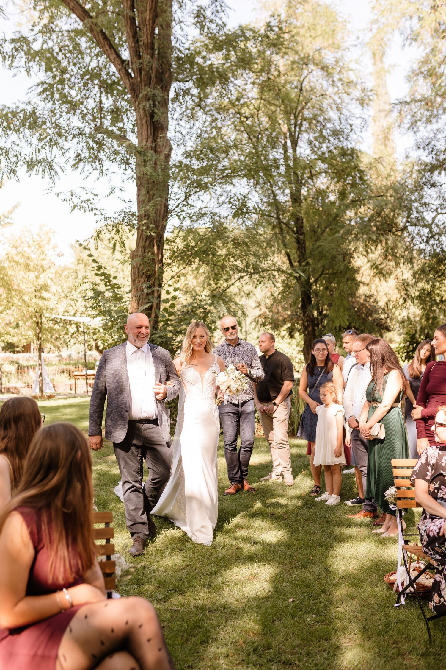 Bride walks down outdoor wedding aisle with escort among guests seated on wooden benches under tall trees.