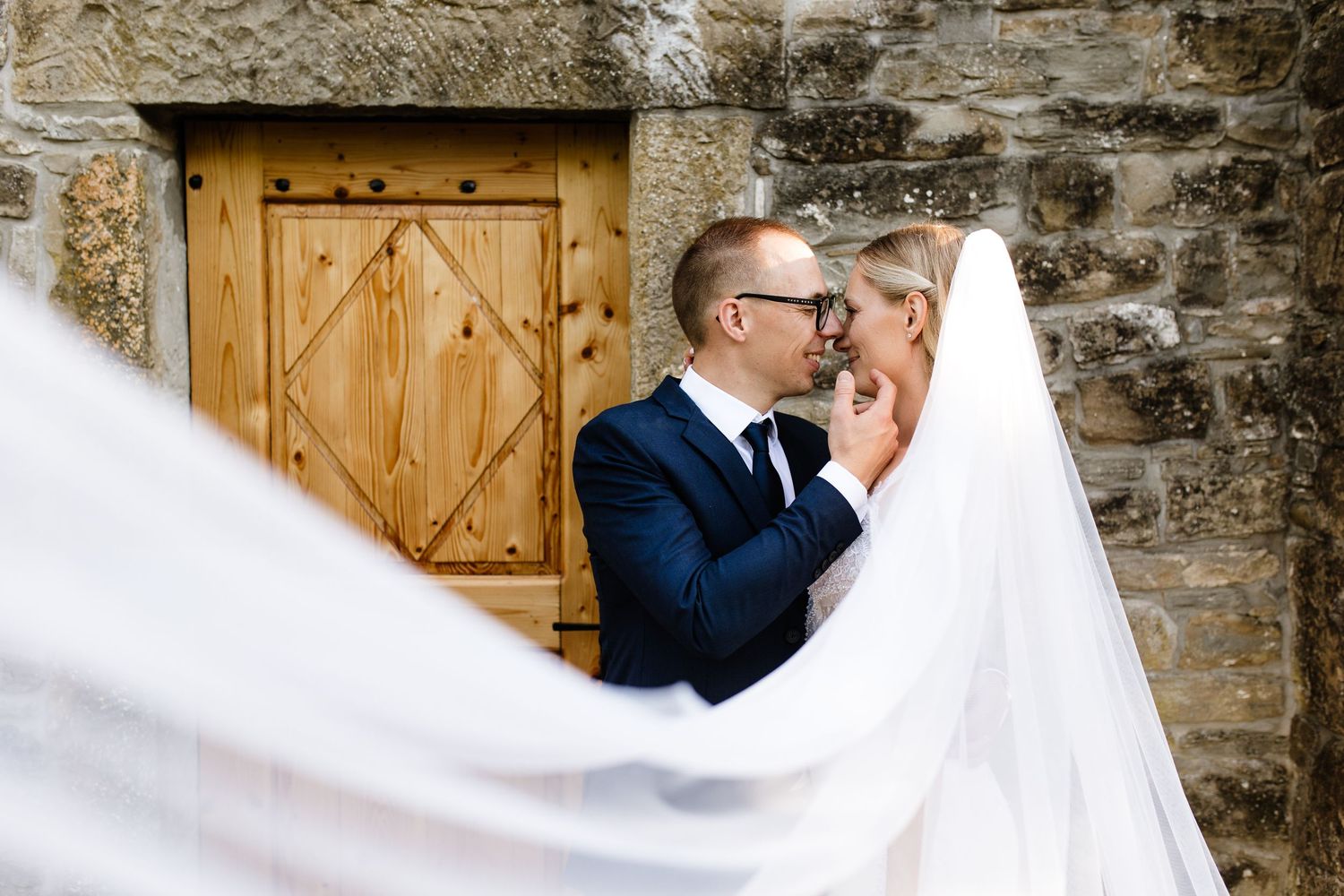 Bride and groom sharing intimate moment wrapped in veil against rustic stone church wall.