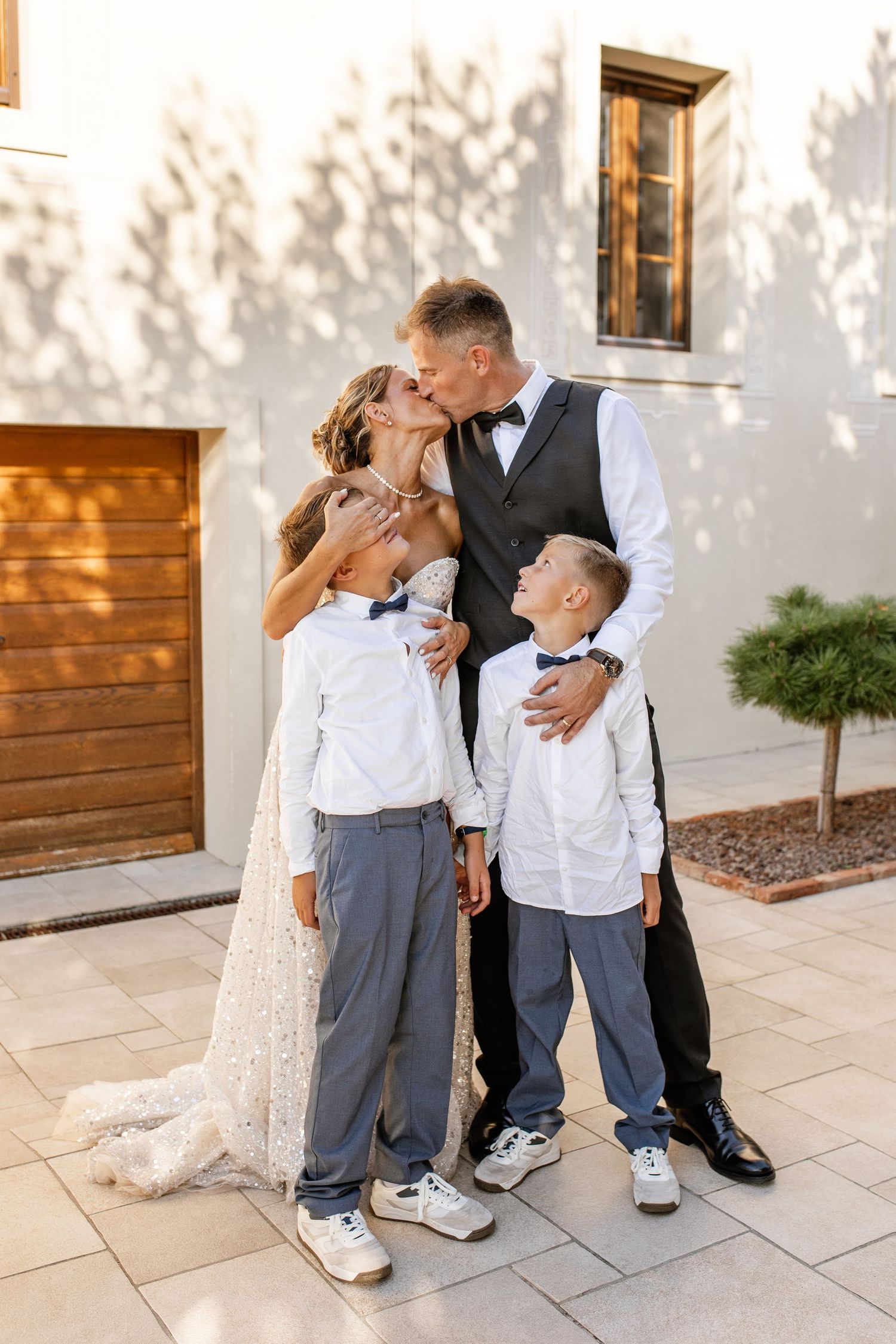 Bride and groom kissing while two young boys in bow ties hug them outside.