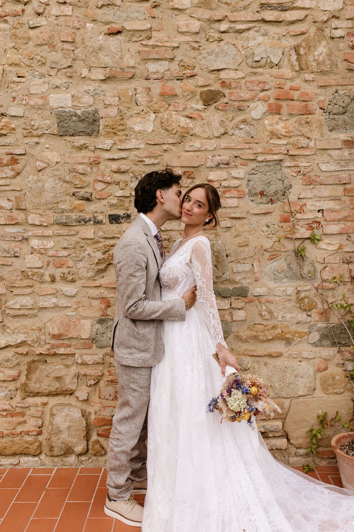 Groom kisses bride on cheek as she smiles holding wildflower bouquet by stone wall.