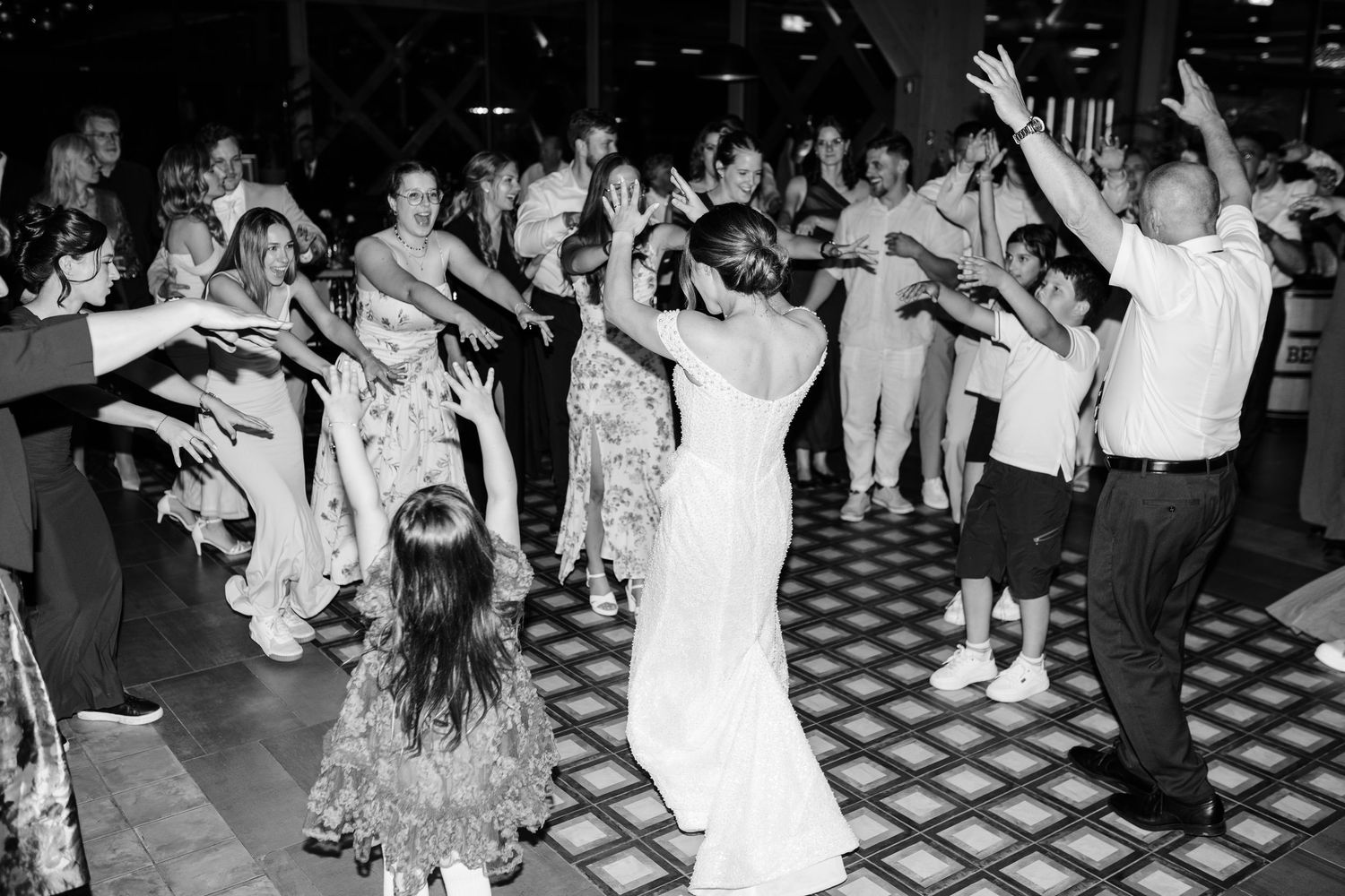 Black and white photo of bride surrounded by cheering wedding guests on crowded dance floor.