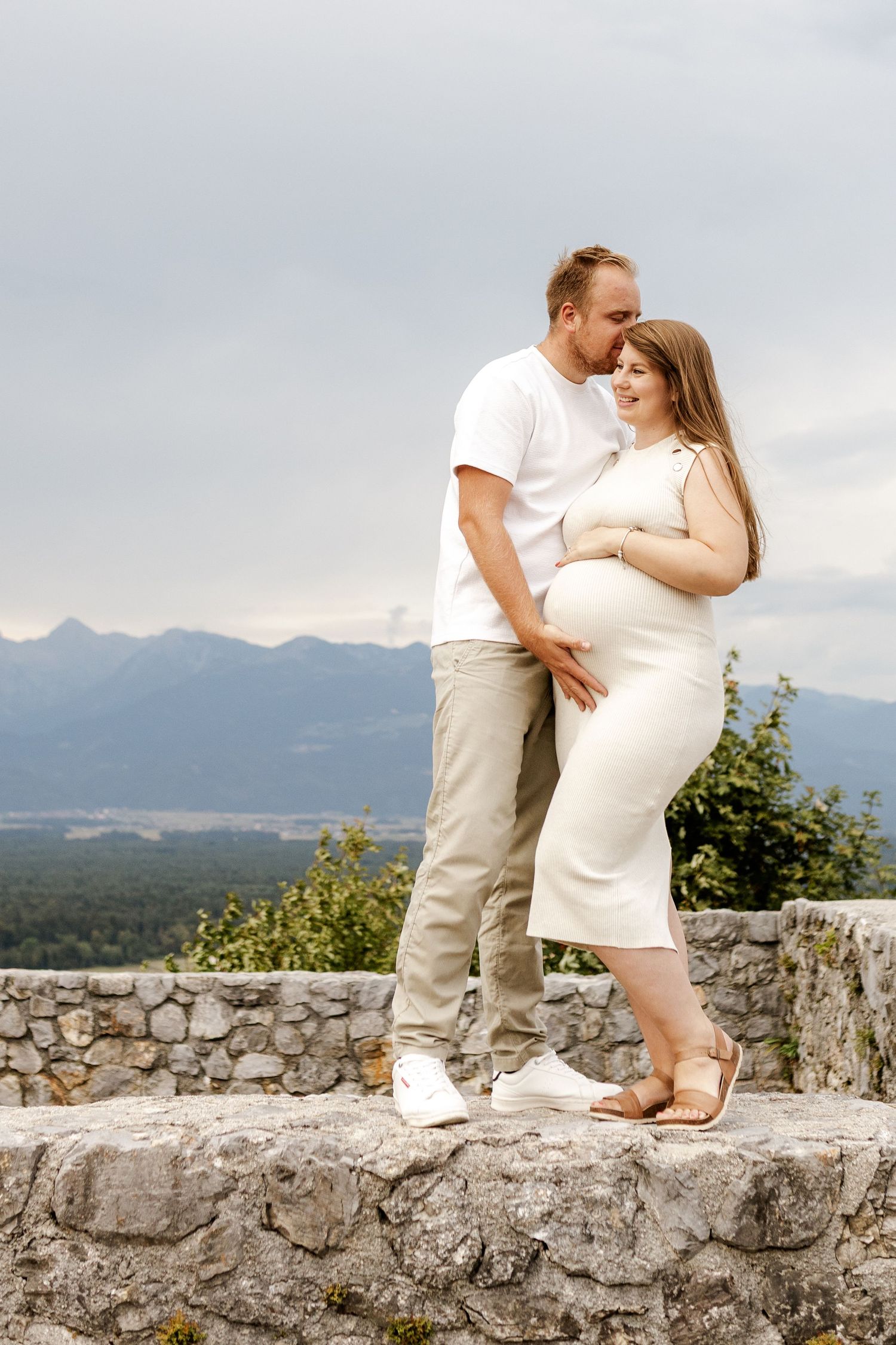 Pregnant couple embracing on stone wall with mountain landscape backdrop, woman in white dress, man in white t-shirt.