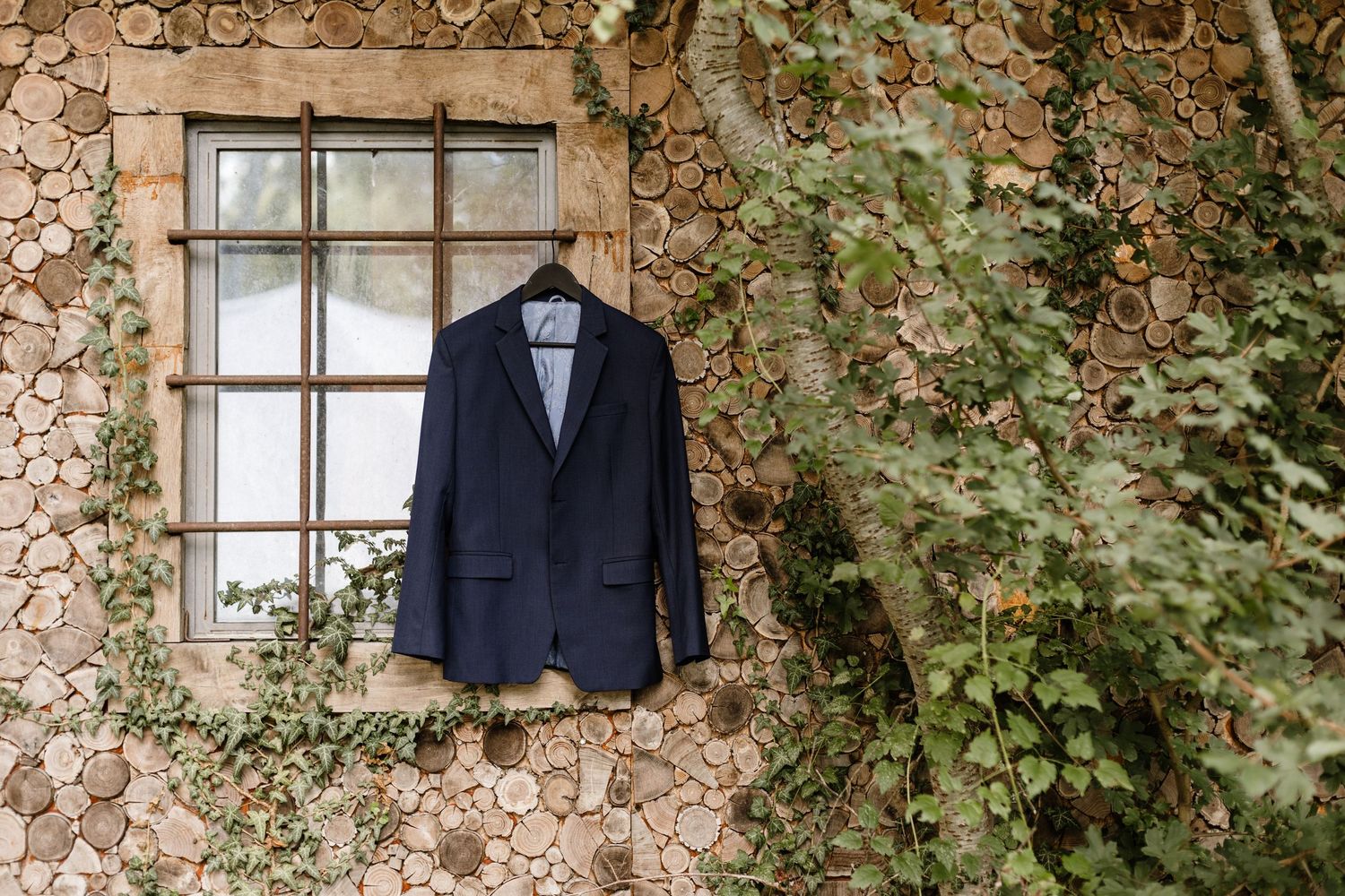 Navy blue suit jacket hanging on a hanger against a rustic stone wall with ivy and a white window.