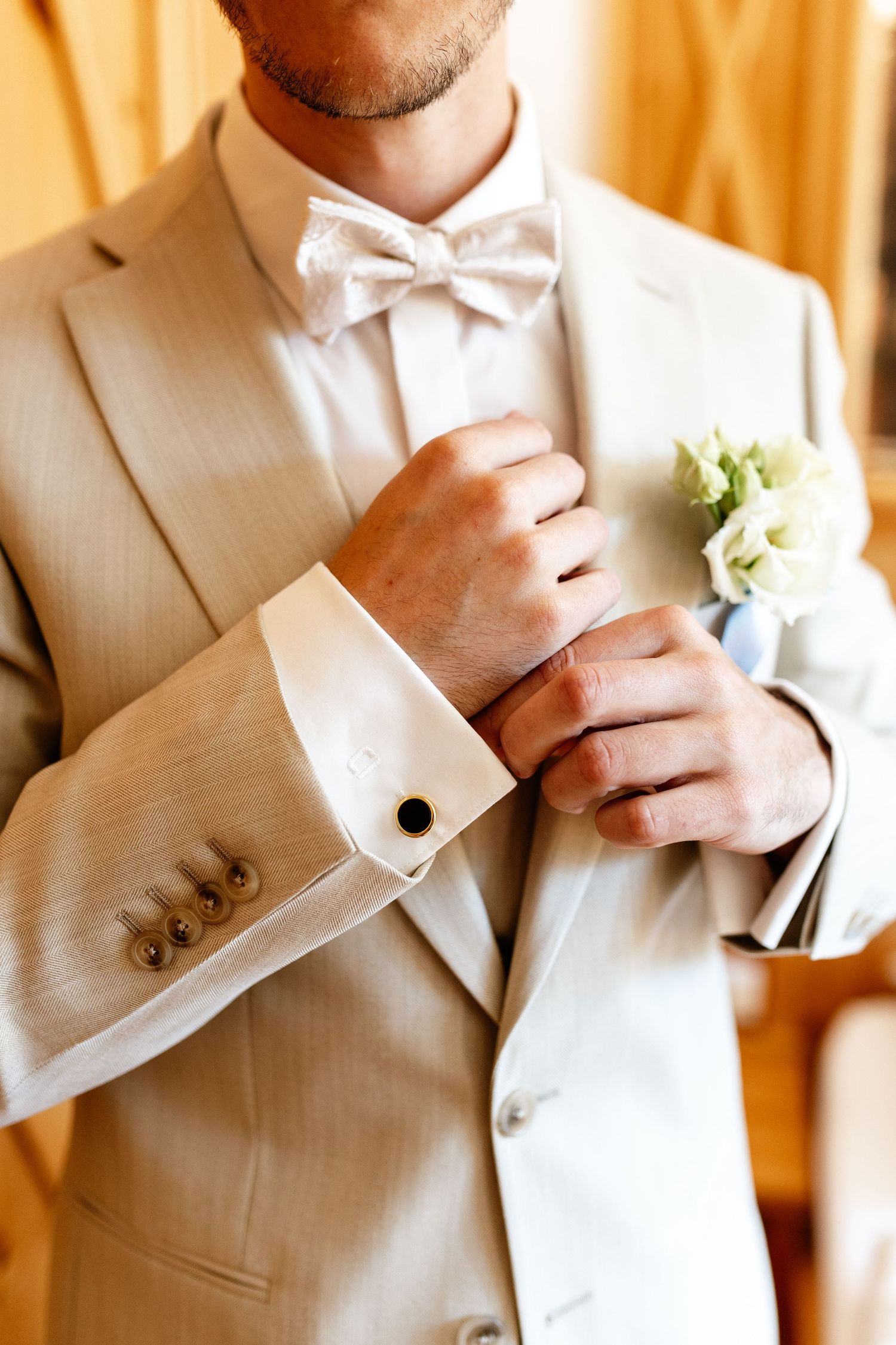 Groom in beige suit adjusting cufflinks, wearing white bow tie and floral boutonniere on wedding day.