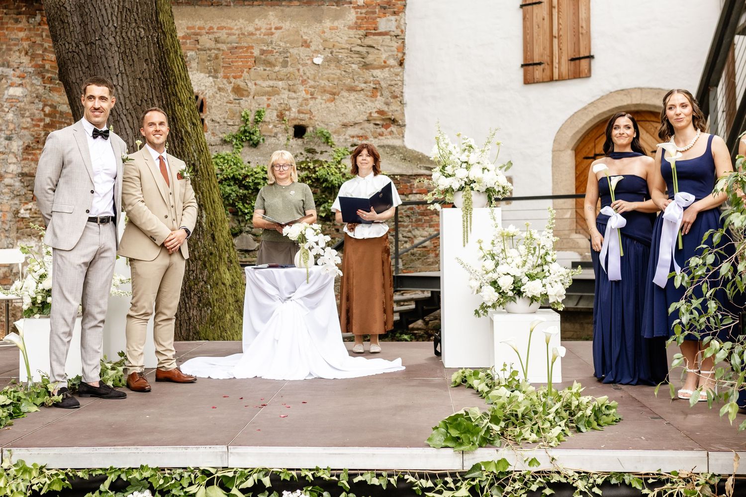 Elegant outdoor wedding ceremony with two grooms, officiant, and bridesmaids in navy dresses at a rustic Italian venue.