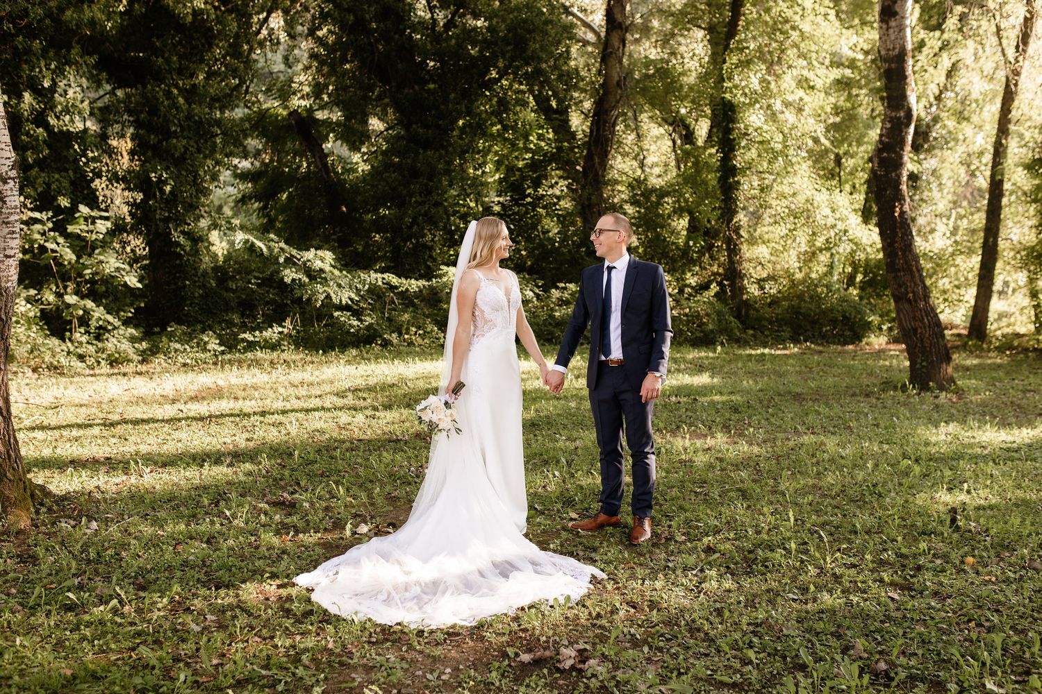 Bride and groom holding hands in a lush green forest setting, bride wearing a flowing white gown.