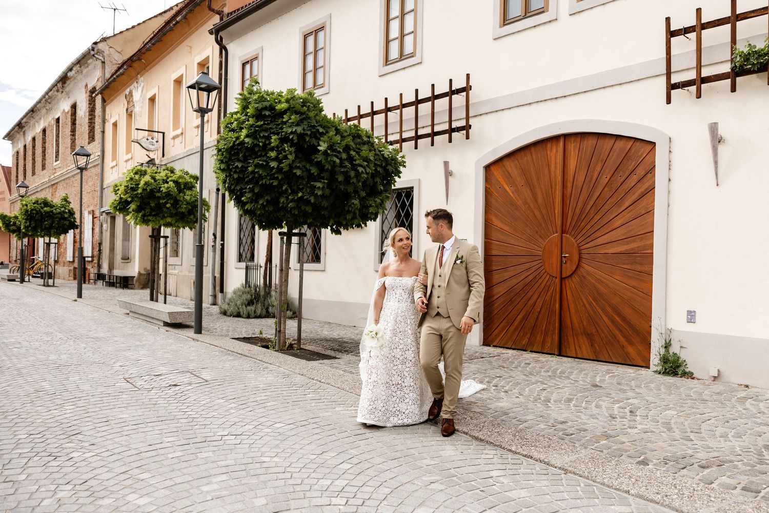 Bride and groom walking hand in hand on a cobblestone street lined with trees and historic European buildings.