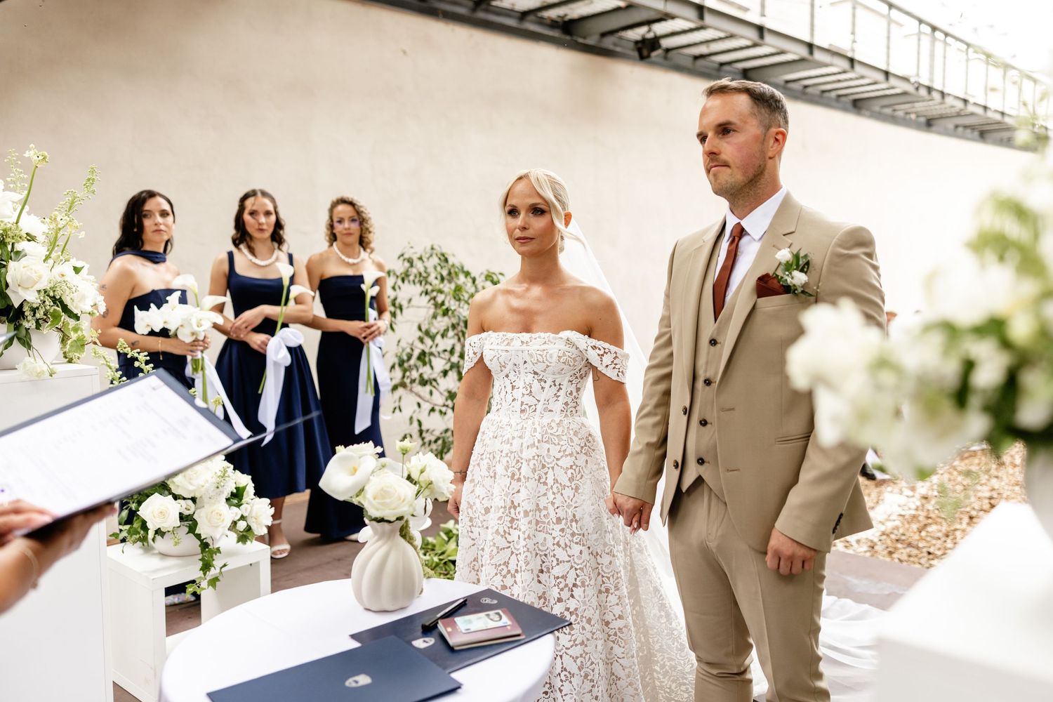 Bride in lace gown and groom in tan suit hold hands at outdoor wedding ceremony with bridesmaids in navy dresses.