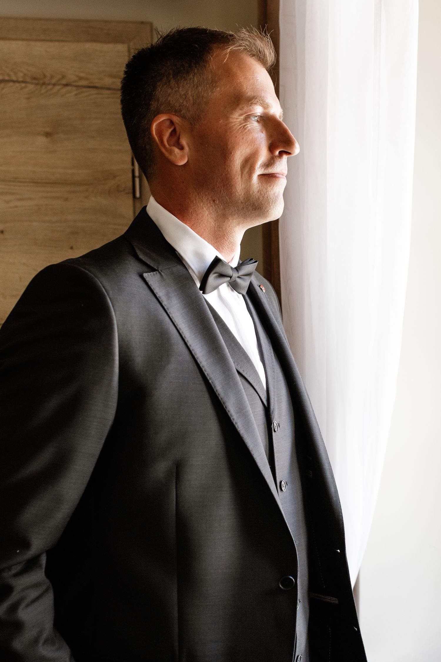 A groom in a dark gray suit with bow tie gazes thoughtfully out a window in soft natural light.