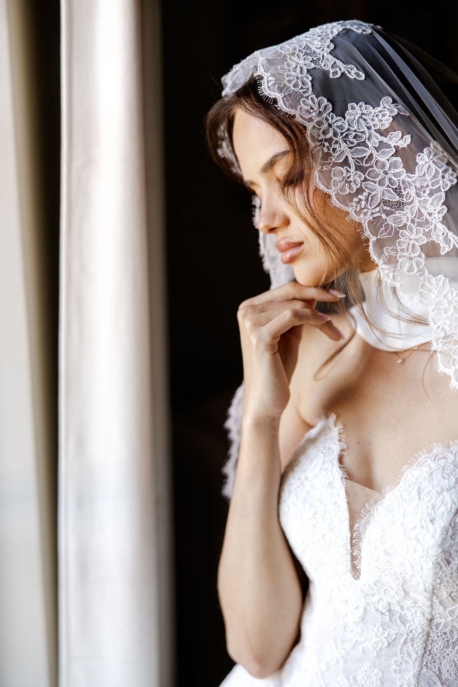 Bride in lace wedding dress and veil gazing down by window light.