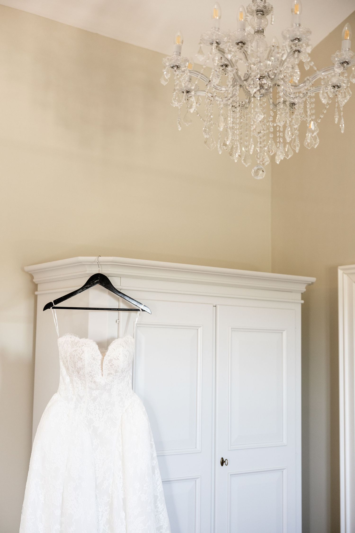 Elegant white lace wedding dress hanging on a black hanger over a white armoire beneath a crystal chandelier.