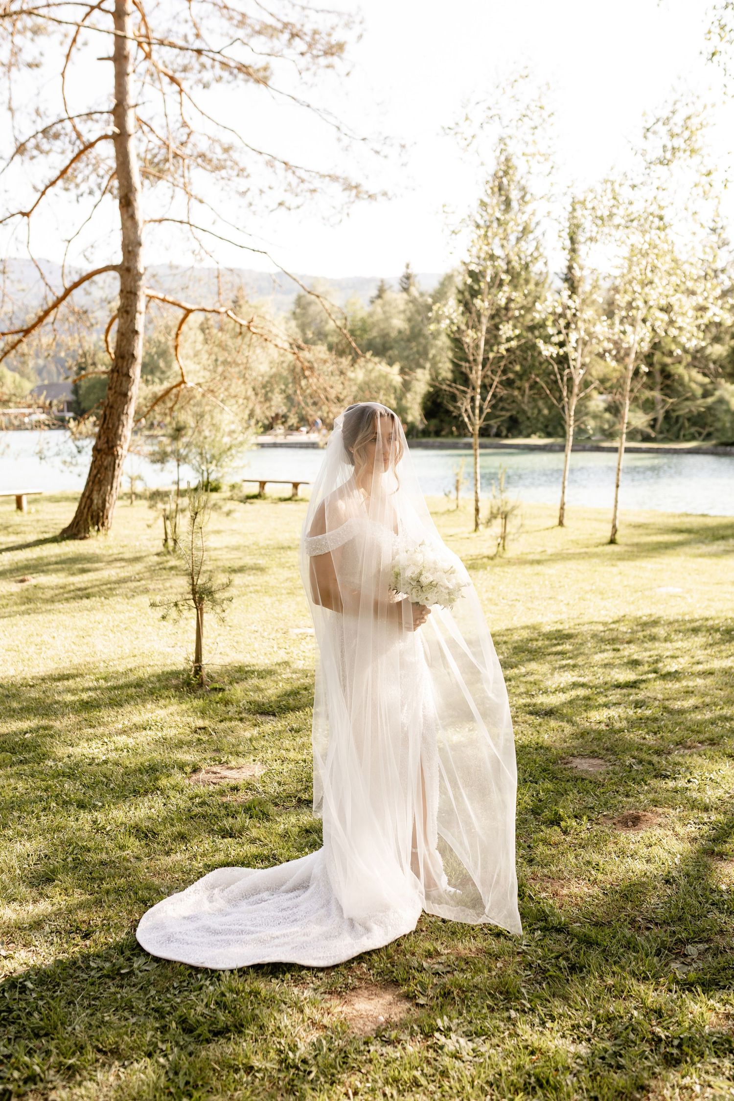 Bride in a flowing white wedding dress and veil standing in a sunlit garden near a serene lake.