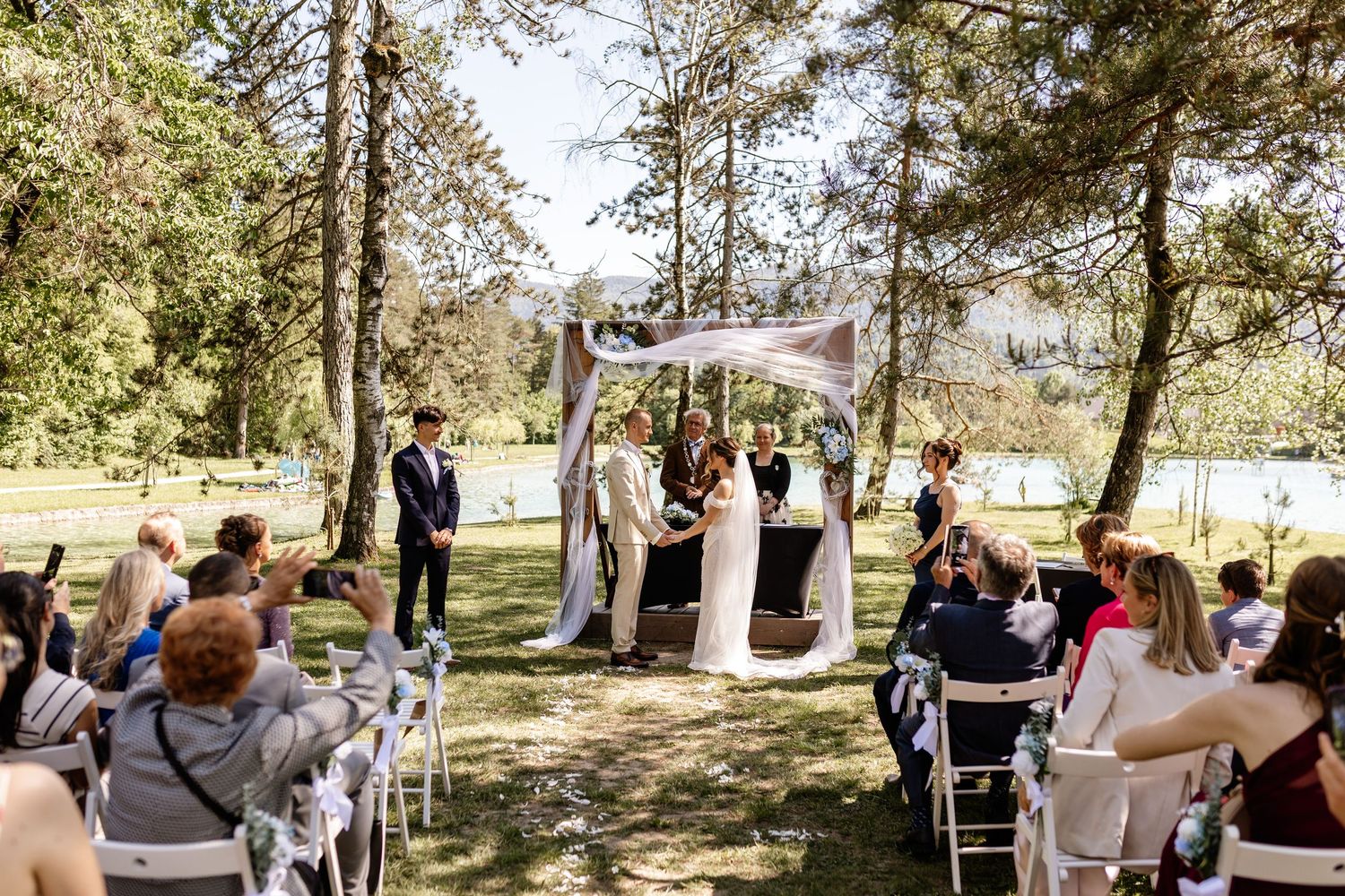 Outdoor wedding ceremony under trees with couple at floral arch, officiant, and seated guests on white chairs.