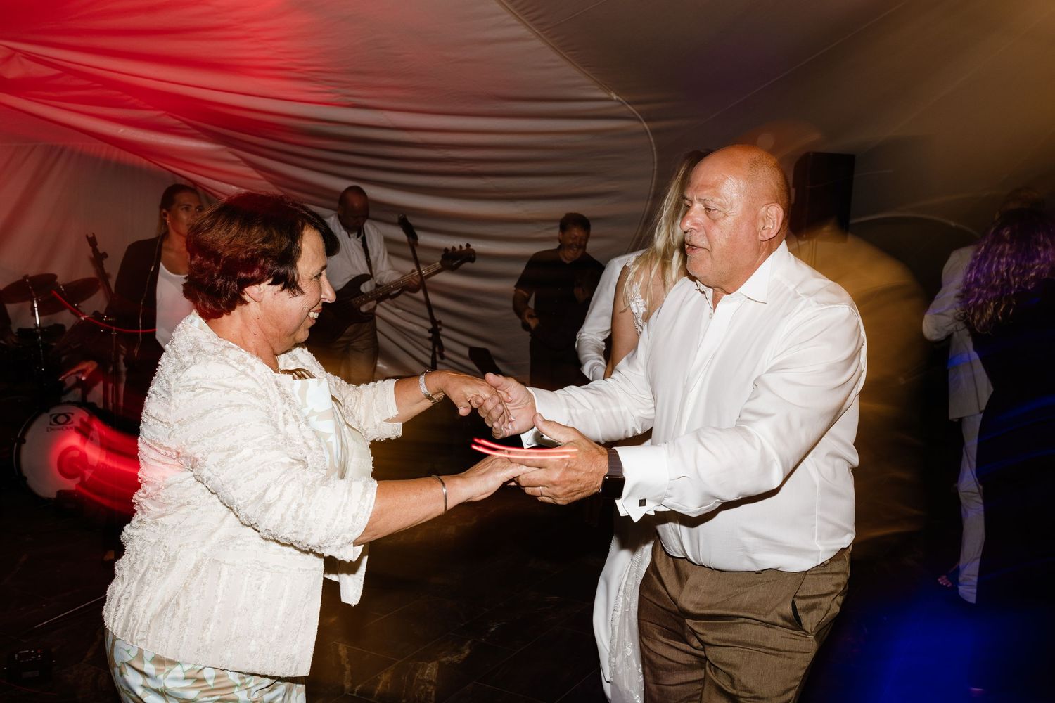 Couple dancing together at an outdoor evening event under a white tent with a live band performing in the background.