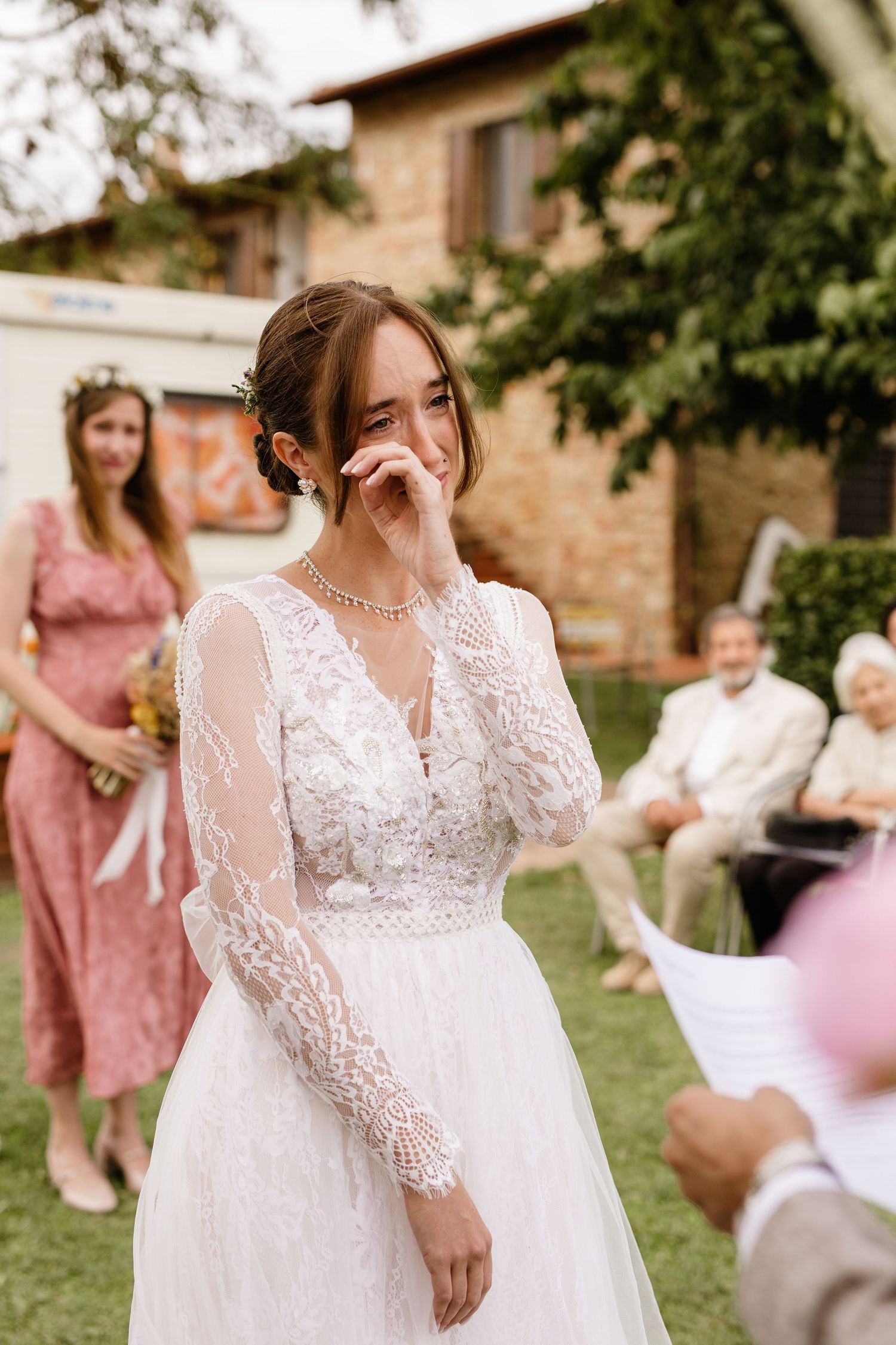 Emotional bride in lace wedding gown wipes tear during outdoor ceremony in Tuscany.